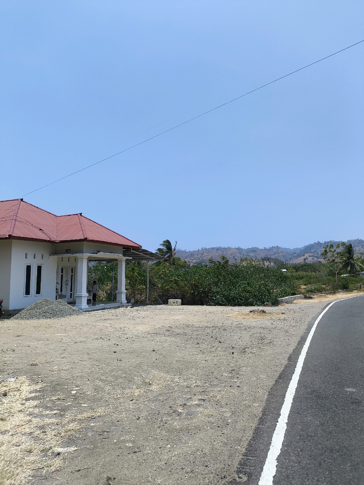 In Kwandang, Indonesia, a house with a red-tiled roof and white walls sits on a lot adjacent to a paved road with a white dividing line. A pile of gravel is located to the left of the house. A person stands on the porch of the house. The house is surrounded by dry, dusty land with some vegetation. In the background, a range of dry, sparsely vegetated hills rises under a clear blue sky. A utility line stretches across the sky from left to right.
