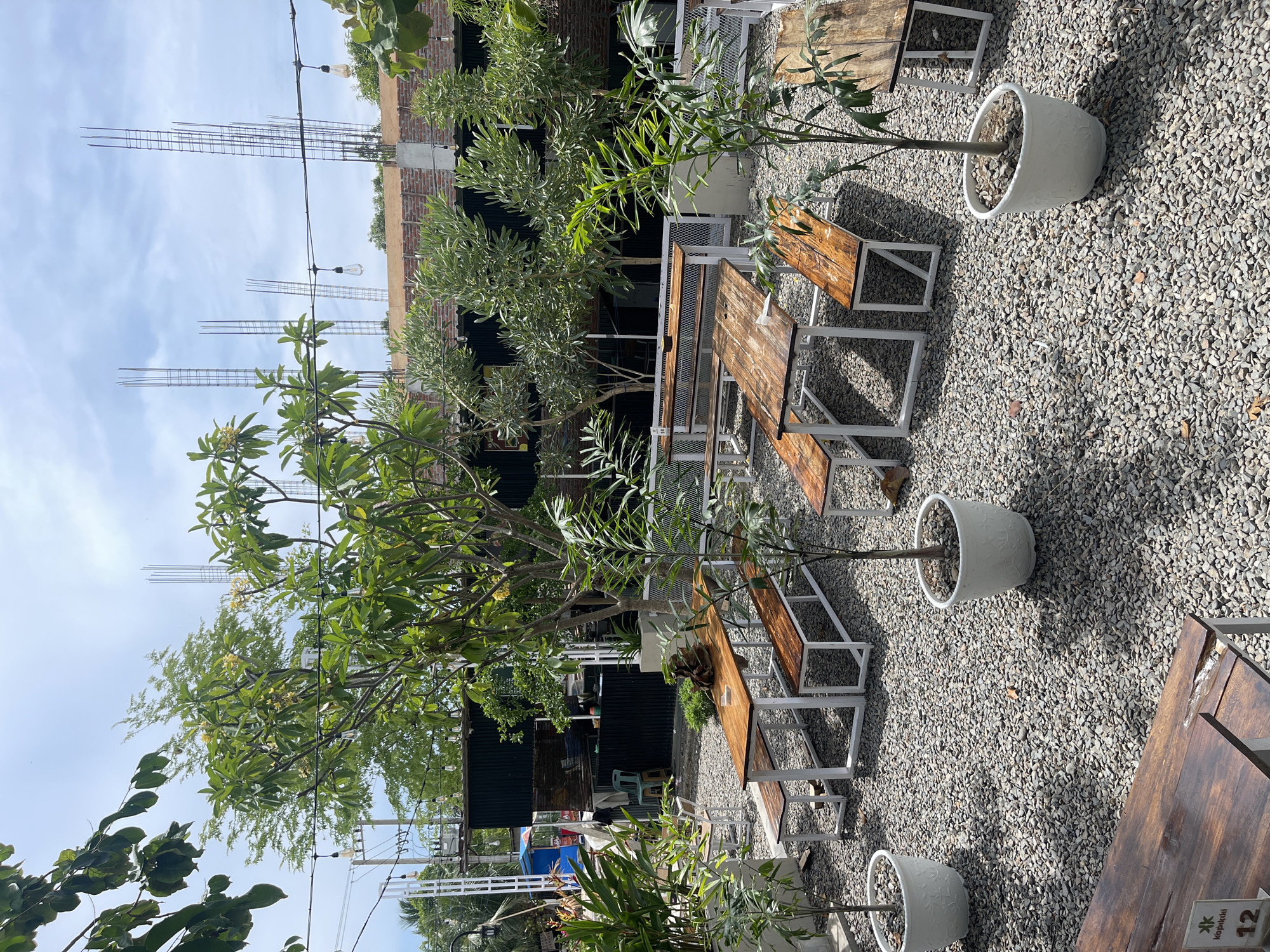 This image depicts an outdoor seating area at a cafe or restaurant in Pulokiton, Indonesia. The ground is covered with small, grey gravel. Several wooden picnic tables with white metal frames are arranged on the gravel. Some tables are empty, while others have benches that are unoccupied. Small trees and potted plants are interspersed throughout the seating area. In the background, a building under construction is visible, with exposed rebar and brickwork. String lights with visible bulbs hang above the seating area, and there are power lines and poles in the distance. A black corrugated metal structure is part of the building's facade. The sky is partly cloudy. There are no people or animals clearly visible in the frame.