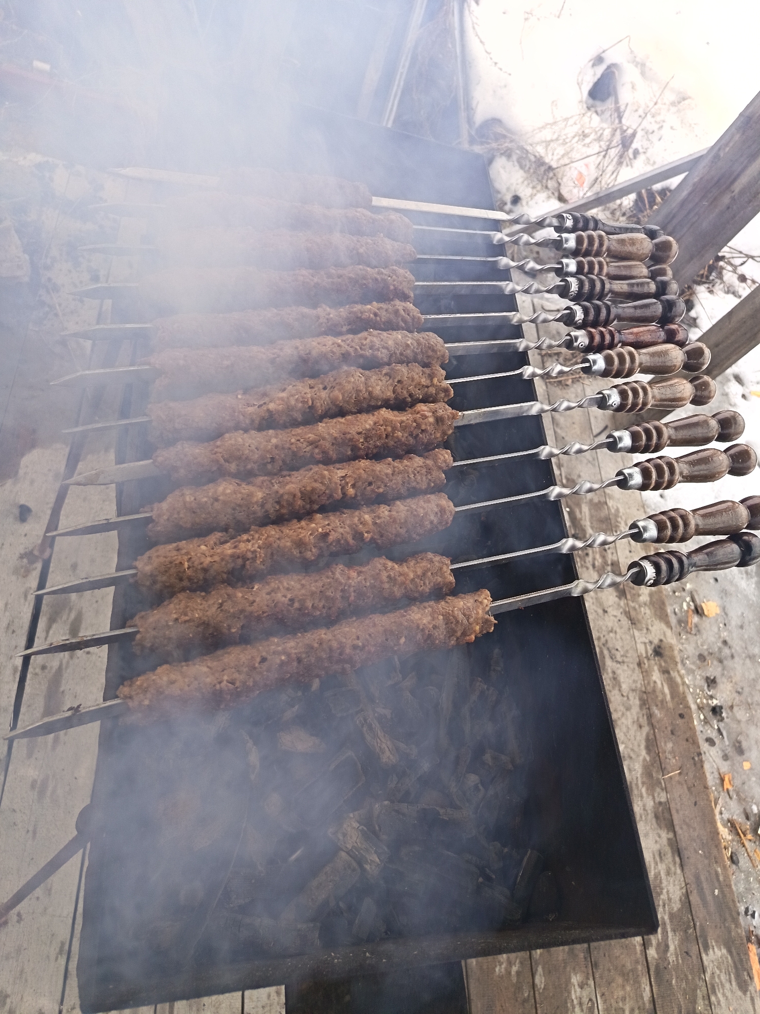 The image shows several skewers of what appears to be meat kebabs cooking over a grill, likely outdoors. Smoke is rising from the grill, partially obscuring the kebabs. The skewers are arranged horizontally across the grill, each holding a long roll of meat. The handles of the skewers are visible on the right side, featuring a dark brown and silver color scheme. The grill is situated in what seems to be a wooden structure or deck. Outside the deck, hints of foliage and what may be a building or structure can be seen. There is no information to support that this picture was taken in Nizhnevartovsk, Russia.