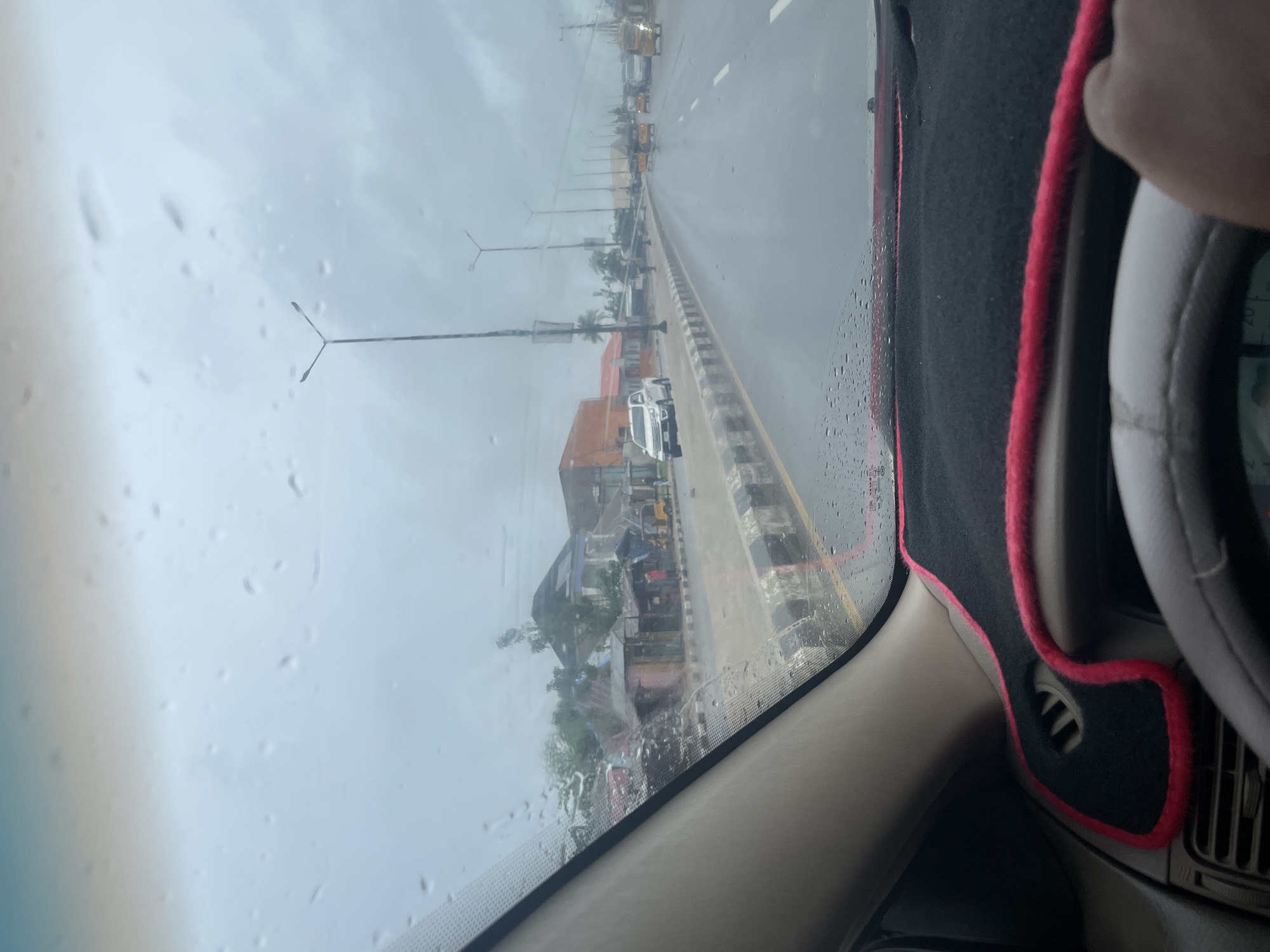The image shows a view from inside a car in Jalingo, Nigeria, looking out through a rain-streaked windshield at a street scene. The sky is overcast, and the road and surroundings appear wet, indicating it is raining or has recently rained.
On the left side of the road, there are several buildings, some of which appear to be small shops or market stalls. One building has an orange facade. There are various vehicles on the road, including a white pickup truck, several yellow motorized rickshaws (locally known as "Keke Napep"), and other cars and vans. Some of these vehicles are in motion.
The road itself is paved and has lane markings. There are streetlights visible along the side of the road. In the foreground, the dashboard of the car is visible, with a black dash mat trimmed in pink. A hand is visible on the right side of the steering wheel. The overall impression is of a moderately busy street scene in a Nigerian city during a rainy day.