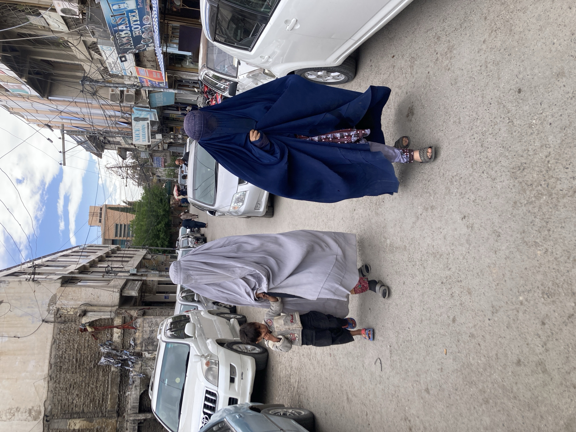Two women wearing burqas, one blue and one grey, walk on a street in Quetta City Tehsil, Pakistan. A child in dark trousers and a light-colored jacket walks alongside the woman in the grey burqa. The street is lined with parked cars and buildings. Overhead, numerous power lines cross the sky. Signs for "Abbasi Hotel" and "Nice Tailors" are visible on the buildings. The sky is partly cloudy.