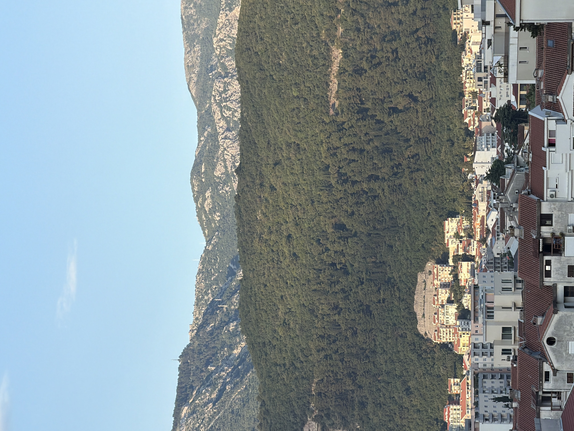 The image captures a panorama of Budva, Montenegro, with a focus on the layered landscape of mountains and urban development. In the foreground, a dense cluster of residential buildings with red-tiled roofs dominates the scene. These structures vary in height and architectural style, suggesting a mix of older and newer constructions. Moving towards the middle ground, the buildings become larger and more modern, with light-colored facades and multiple stories. A prominent hill, partially cleared and terraced with buildings, rises behind this urban sprawl. This hill is topped with a prominent, light-colored structure.
Behind the urban areas, a vast, densely forested mountain slope stretches across the mid-ground. The trees are predominantly a deep green, creating a thick canopy that obscures much of the underlying terrain. Further in the background, a second, more rugged mountain range rises. These mountains appear rocky and less vegetated, with lighter tones that contrast with the dark forest. A few communication towers and utility poles are visible on the slopes of these distant mountains, hinting at infrastructure.
The sky above is a clear, pale blue, with a few wispy clouds scattered near the horizon. The lighting suggests it is late afternoon or early evening, with the sun casting a warm glow on the buildings and highlighting the contours of the landscape. No people are visible in the image, and the scene is devoid of any overt action or interaction. The overall impression is one of a coastal city nestled between the sea and mountainous terrain.