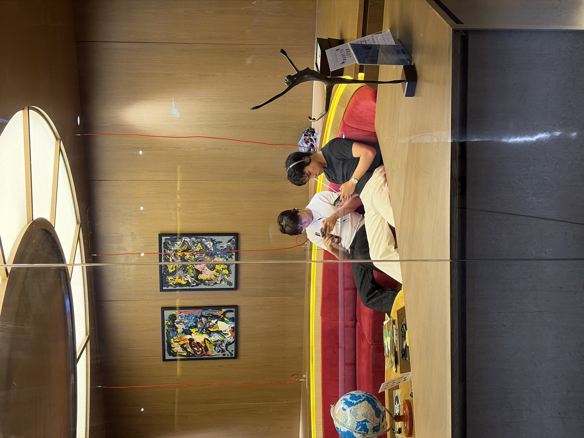 Two people are seated on a red cushioned bench, interacting with mobile phones. One person wears a black shirt and headphones, while the other wears a white shirt. Artwork hangs on a wood-paneled wall, with a unique lighting fixture to the left. A modern sculpture stands on a wooden surface near the people. A globe rests near the edge of the bench, and informational signs are present. The location is likely a public space in Bangkok, Thailand.