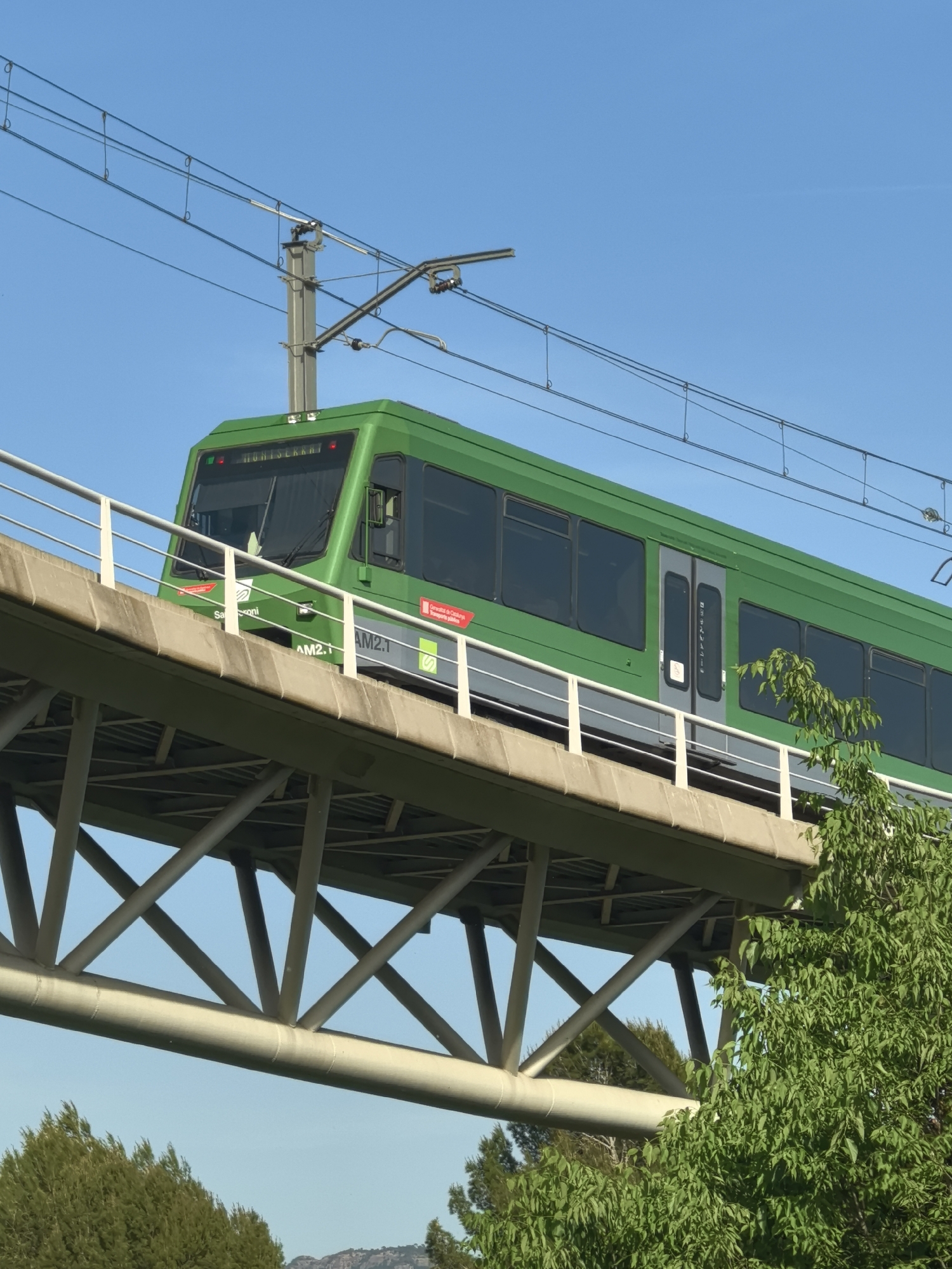 The image displays a vibrant green train moving along an elevated track under a clear blue sky. The train, a modern electric multiple unit, is the primary subject, featuring multiple windows and doors. On its front, an illuminated destination sign prominently reads "MONTSERRAT". Below this, text including "AM2.T" and a logo (possibly "Sargassoni") are visible. Further along the side, near a door, a red logo with "Generalitat de Catalunya" and "Transportes públicos" (public transport) is present, alongside another "AM2.T" and the S-logo. Through the train's front window, text on an interior door can be seen, stating "Porta automàtica" and "Prem per obrir" (Catalan for "Automatic door" and "Press to open").
The train is situated on an elevated concrete platform supported by a sturdy metal truss bridge structure, painted in a light, neutral color. Grey railings line the edges of the track. Above the train, a complex network of overhead power lines (catenary system) is visible, indicating an electric railway.
Lush green trees and foliage frame the lower part of the image, with their leaves contrasting against the blue sky and the bridge's structure. In the distant background, subtle outlines of mountains or hills can be discerned. The bright and cloudless blue sky suggests a clear and sunny day, likely morning or afternoon. The scene depicts a functional transportation system harmoniously integrated into a scenic natural environment, specifically in Monistrol de Montserrat, Spain.