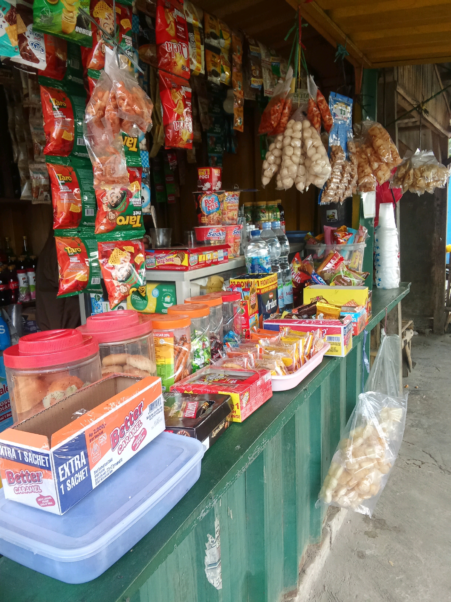 The image shows a small, outdoor food stall in Warunggunung, Indonesia. The stall is laden with a variety of packaged snacks, drinks, and food items. Prominently displayed on the counter are several plastic containers filled with what appear to be baked goods, possibly bread or pastries. Various bags of chips and snacks hang from the ceiling of the stall. In the foreground, a box labeled "Better Caramel" is positioned on top of a blue plastic container. A large plastic bag filled with puffed snacks hangs from the side of the stall. The overall scene depicts a typical informal market setting, with an abundance of goods for sale.