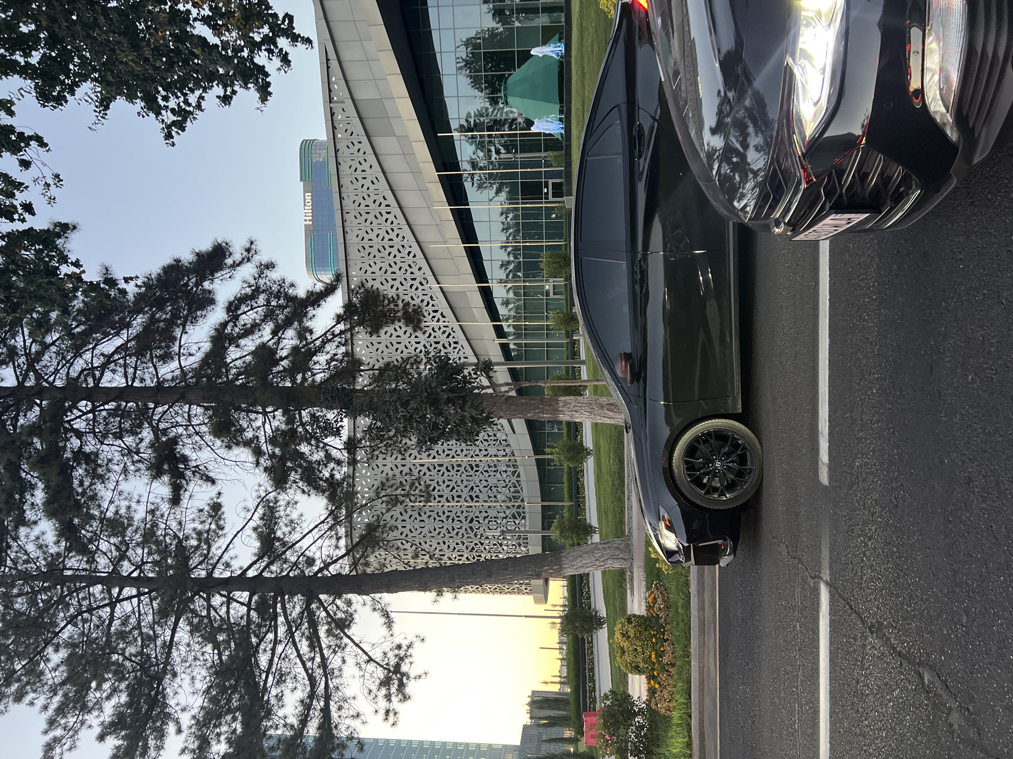 This image depicts a street scene in Tashkent, Uzbekistan, likely near the Humo Arena or the nearby Hilton hotel, given the presence of the Hilton signage on a modern building. In the foreground, two black sedans are driving on a paved road with white lane markings. The car on the right is closer to the camera, showing its front grill, headlights, and a license plate that is partially visible and appears to have Cyrillic characters. The car on the left is further down the road, showing its side profile and black wheels.
Behind the road, a landscaped median separates it from a large, modern building with a distinctive geometric patterned facade and large glass windows. Trees, including tall pine trees, are present in the foreground and along the median, casting shadows. The building's architecture suggests a contemporary design. In the distance, another tall building is visible, and the sky suggests it is either early morning or late afternoon, with soft light. Green decorative elements, possibly a tent-like structure and fountains with blue lighting, are visible on the lawn in front of the large building. The overall scene conveys a sense of urban development and modern infrastructure.