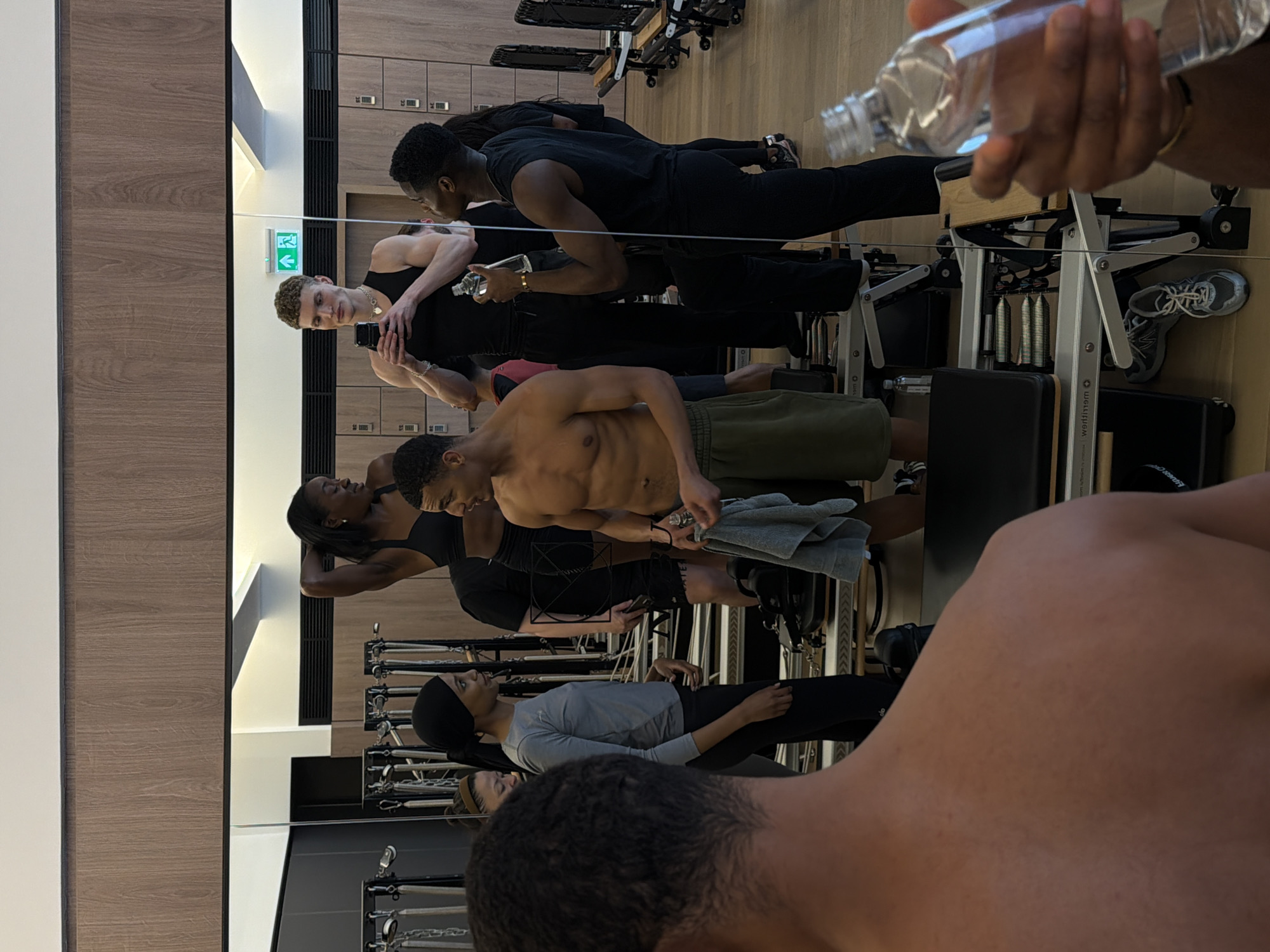 The image captures a group of people in what appears to be a pilates or fitness studio. Several individuals are positioned on pilates reformer machines, while others stand and observe. The machines are characterized by their sliding carriages, springs, and straps. The studio environment features a wall-mounted mirror, and a wall with storage lockers. The specific location and address in London, United Kingdom, are not discernible from the image.