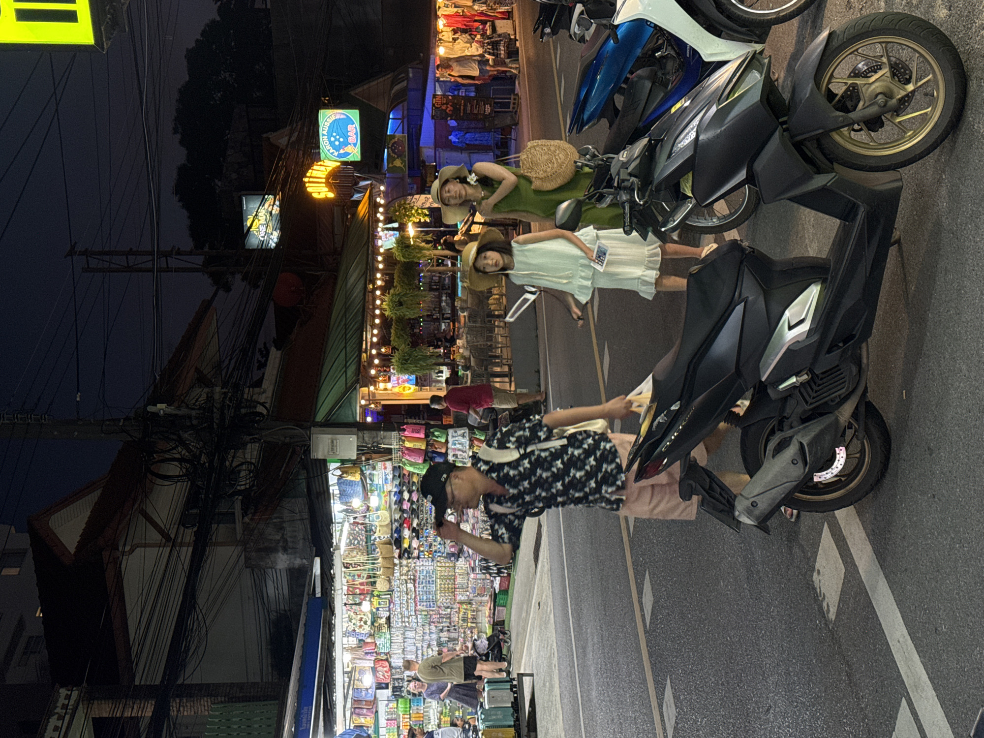 In Karon, Thailand, at night, a street scene unfolds with commercial establishments lining one side of a road. The foreground is dominated by a dark-colored scooter parked on the asphalt. To the right of the scooter, another scooter, primarily blue and white, is partially visible.
A man wearing a patterned short-sleeved shirt, a black baseball cap, and light pink shorts walks towards the right, adjusting his cap. He carries a light-colored bag over his shoulder and another bag in his hand.
Further down the street, a woman in a green dress and a wide-brimmed hat stands with a straw bag on her shoulder. Beside her, a young girl in a light green ruffled dress and a straw hat holds a small white object and a phone. Both are facing left.
In the background, a brightly lit shop displays numerous bags and various merchandise. Several people are visible browsing or walking near the shop. Overhead, a dense network of electrical wires crisscrosses the scene. Neon signs and streetlights illuminate the area, casting a warm glow on the surroundings. To the right, a bar with outdoor seating is visible, with patrons seated at tables.