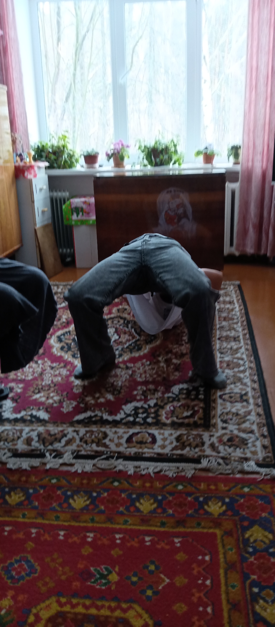 The image shows a person performing a backbend in a room. The person is wearing jeans and a white shirt. The background includes a window with plants on the sill, a cabinet, a radiator, and two rugs on the floor. The location is Unknown, Unknown.