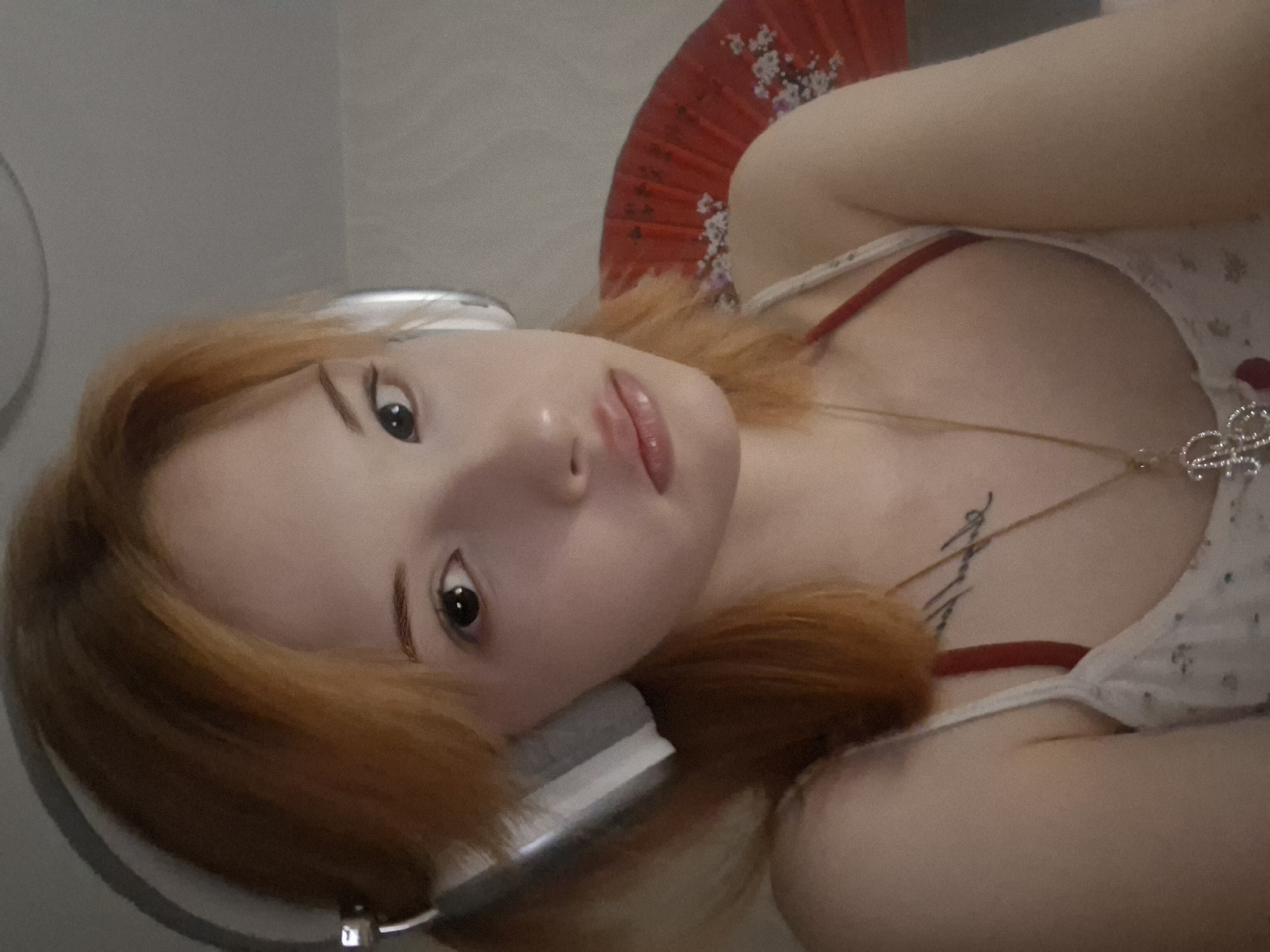 A young woman with light skin and auburn hair is pictured from a slightly angled perspective. She is wearing headphones and a light-colored camisole top with a red strap visible. A necklace with a decorative pendant is around her neck. She has a small tattoo on her chest. Behind her head is a red folding fan. The location is Chaikovsky, Russia.
