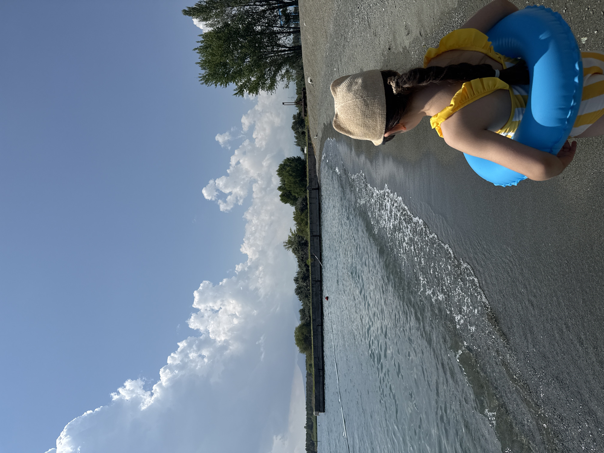 The image shows a young girl wearing a straw hat and a yellow and white striped swimsuit. She has a blue inflatable ring around her body and is resting on a sandy beach. The sea is gently lapping at the shore. In the background, there are trees and a clear blue sky with some fluffy white clouds. The scene appears to be from a sunny day at the beach, possibly during summer. The girl is looking down and appears to be relaxed. The location is Tsamakaberd, Armenia.