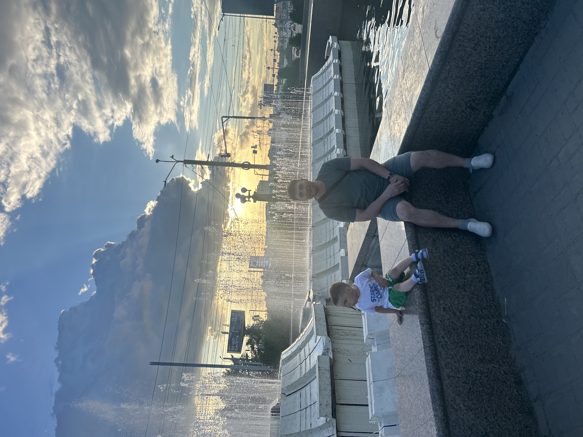 The image depicts a father and son sitting on a granite ledge in front of a large, tiered fountain in Omsk, Russia. The fountain has multiple levels with water jets spraying upwards, creating a misty effect. The son, wearing a white t-shirt and green shorts, is seated to the left of the father. The father, dressed in a grey t-shirt and grey shorts, is to the right. In the background, the setting sun casts a warm glow through dramatic, cloudy skies, with silhouetted buildings and overhead power lines visible. The overall scene is set in an urban environment, likely a public square or park in Omsk.