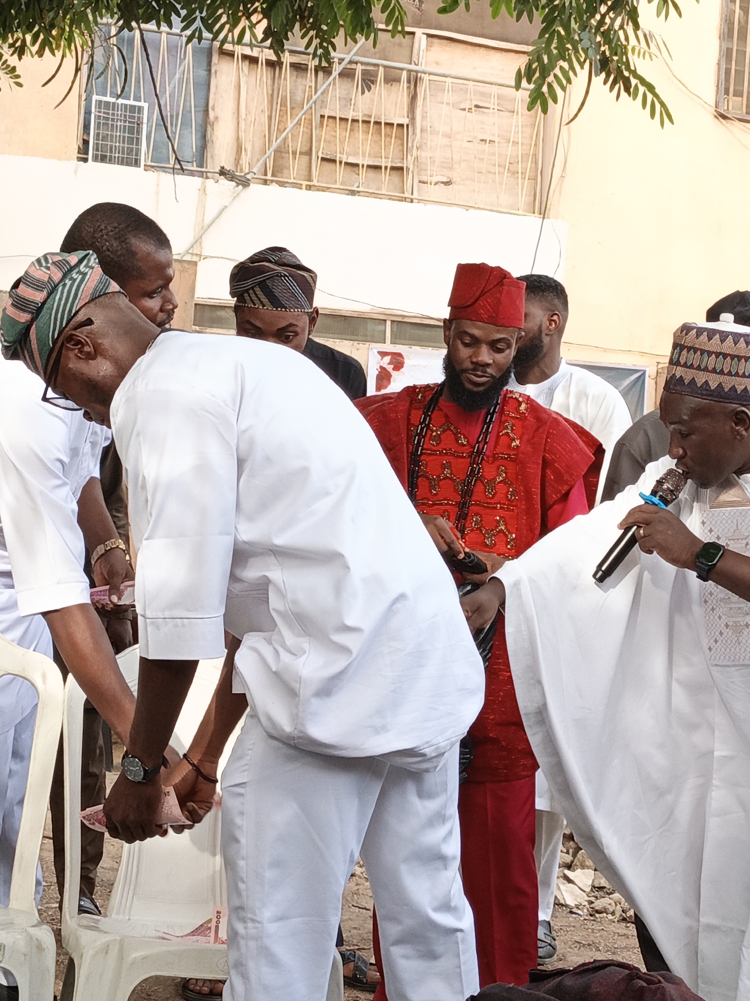 An outdoor gathering of several Nigerian men is depicted in this daylight scene from Gafan, Nigeria. The focus is on a central activity involving money and communication.
In the immediate foreground, to the right, a man in a white traditional robe (possibly an Agbada) and an embroidered cap holds a black microphone to his mouth, seemingly speaking or presiding over the event. He wears a dark smartwatch on his left wrist. Adjacent to him, towards the center, another man dressed in a vibrant red traditional outfit with intricate gold embroidery and a matching red cap looks down intently at the activity unfolding before him. He has a full beard and is adorned with a dark beaded necklace.
The central action involves two men, both dressed in white traditional attire, who are bending forward. The man on the left wears a striped green and white cap, glasses, and a gold wristwatch. He, along with the other man in white, is actively handling a stack of pink banknotes. Several 200 Nigerian Naira notes are clearly visible, with some resting on a white plastic chair. The men appear to be either collecting, distributing, or counting the money during the event.
In the background, other men are partially visible, some wearing traditional caps, observing the proceedings. The setting appears to be an unpaved outdoor area with a light-colored building featuring barred windows in the background. Green foliage hangs from above, providing natural shade. The overall atmosphere appears focused and engaged, consistent with a community gathering or ceremony.