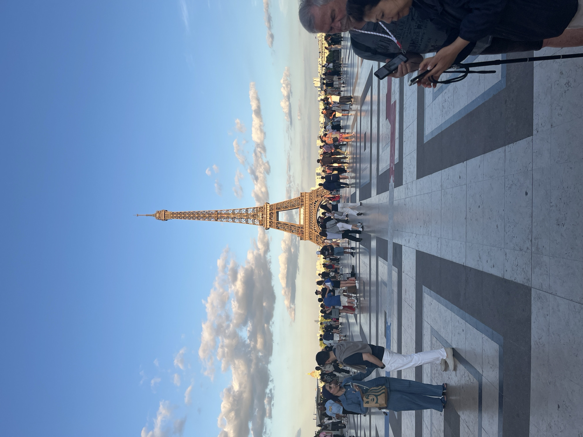 The image depicts a sunny day in Paris, France, with the Eiffel Tower prominently featured in the background. A crowd of tourists are gathered, suggesting a popular tourist attraction. The ground is paved with large, square tiles, arranged in a geometric pattern. Some clouds are visible in the bright blue sky, indicating fair weather. The time of day appears to be late afternoon or early evening, given the warm lighting and the angle of the shadows cast on the pavement. Many people are looking at their phones, possibly taking pictures or navigating. The overall mood seems relaxed and casual, consistent with a tourist experience.