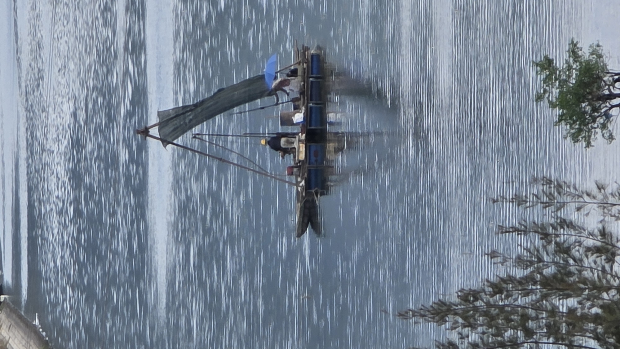 The image shows a boat on the water in Nha Trang, Vietnam. The boat is dark and constructed with a makeshift sail. The boat's occupants appear to be engaged in fishing or a similar water-based activity. Trees and plants are on the right side of the image. The surface of the water reflects light.