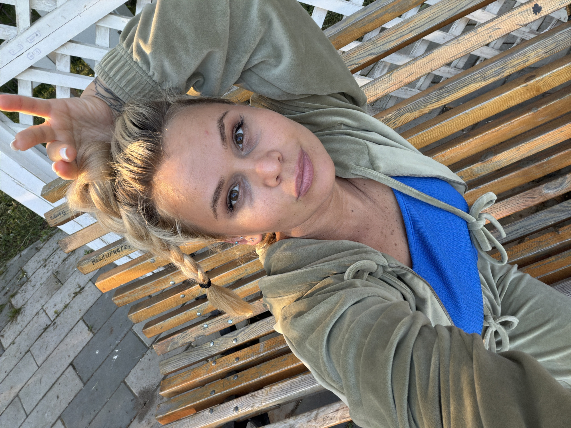 A woman is lying on a wooden park bench. She has blonde hair in pigtails and is wearing a green zip-up jacket over a blue top. She is looking at the camera with her left hand raised. The bench is located near a white lattice fence and a stone walkway. The location could potentially be Sarapul, Russia.