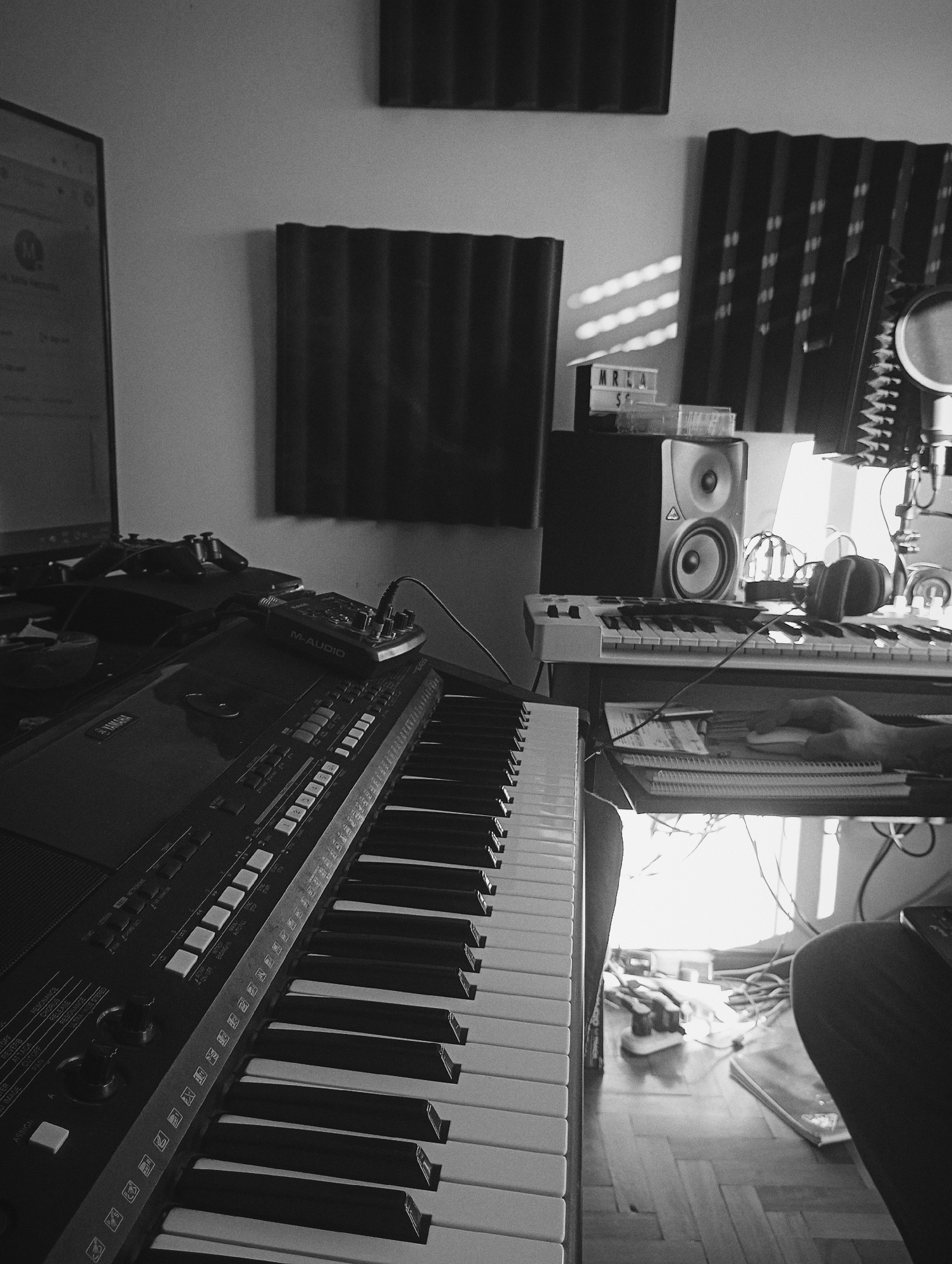This black and white photograph captures a home music studio setup in Buenos Aires, Argentina, bathed in strong, directional sunlight that indicates either late afternoon or early morning. The scene is dominated by musical equipment, suggesting an active creative space.
In the foreground, a large Yamaha keyboard extends from the lower left towards the center, with various controls and keys visible. Text on this keyboard includes "YAMAHA", "REVERB", "CUTOFF", "CHORUS", "RESONANCE", "RELEASE", "ASSIGN", "ATTACK", "OFF", "IMAGE", and "ER". Perched on top of the keyboard is an "M-AUDIO" branded audio interface.
Further back, a person's right hand is visible, operating a computer mouse on a stack of notebooks and papers. Adjacent to this, another keyboard/MIDI controller is situated beneath two studio monitor speakers. Headphones rest on this second keyboard, and a microphone on a stand is positioned to the right. Acoustic foam panels are affixed to the wall, and a small lightbox displaying "MREA" and "S" sits between the speakers. A computer monitor is partially visible on the far left. Gaming controllers are casually placed on the back keyboard. The floor features a wooden parquet, and bright sunlight streams in from a window area, illuminating numerous cables and power strips. The image conveys an atmosphere of creative work and music production.