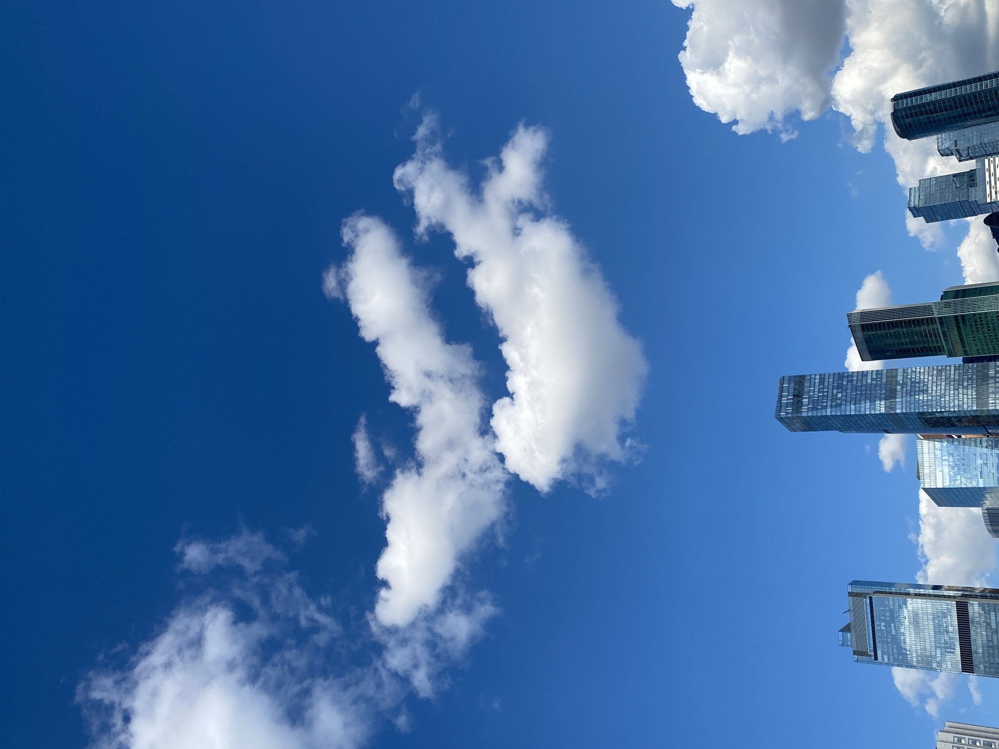 The image shows a low-angle view of a cluster of modern skyscrapers against a vibrant blue sky dotted with white, fluffy clouds. The buildings are sleek and tall, featuring reflective glass facades that mirror the sky and clouds. The sunlight suggests it is daytime. The composition emphasizes the height and scale of the architecture against the expansive sky. The scene is devoid of people or any explicit signs of activity beyond the static presence of the buildings.