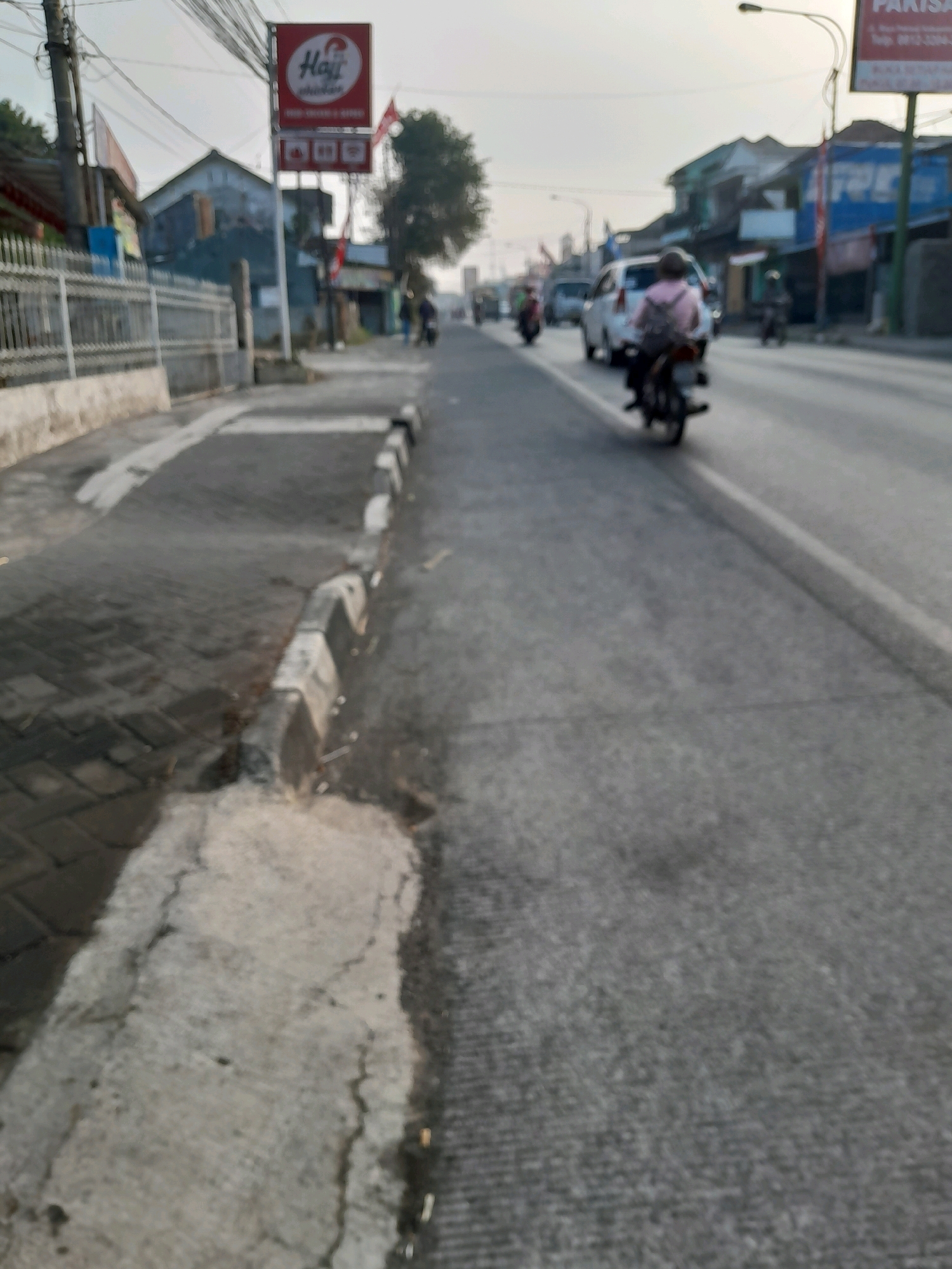The image shows a street scene in Karangpandan, Indonesia, with a focus on a sidewalk and the adjacent road. On the sidewalk, there is a red and white sign with the word "Haji" and a chicken logo. The sidewalk is paved with interlocking bricks in some areas and concrete in others, with a curb separating it from the road. The road is asphalt, and there are vehicles, including motorcycles and a white car, moving along it. One motorcyclist, wearing a pink shirt, is visible in the foreground. Buildings line the side of the road, and overhead wires are present. The overall impression is of a busy urban environment.