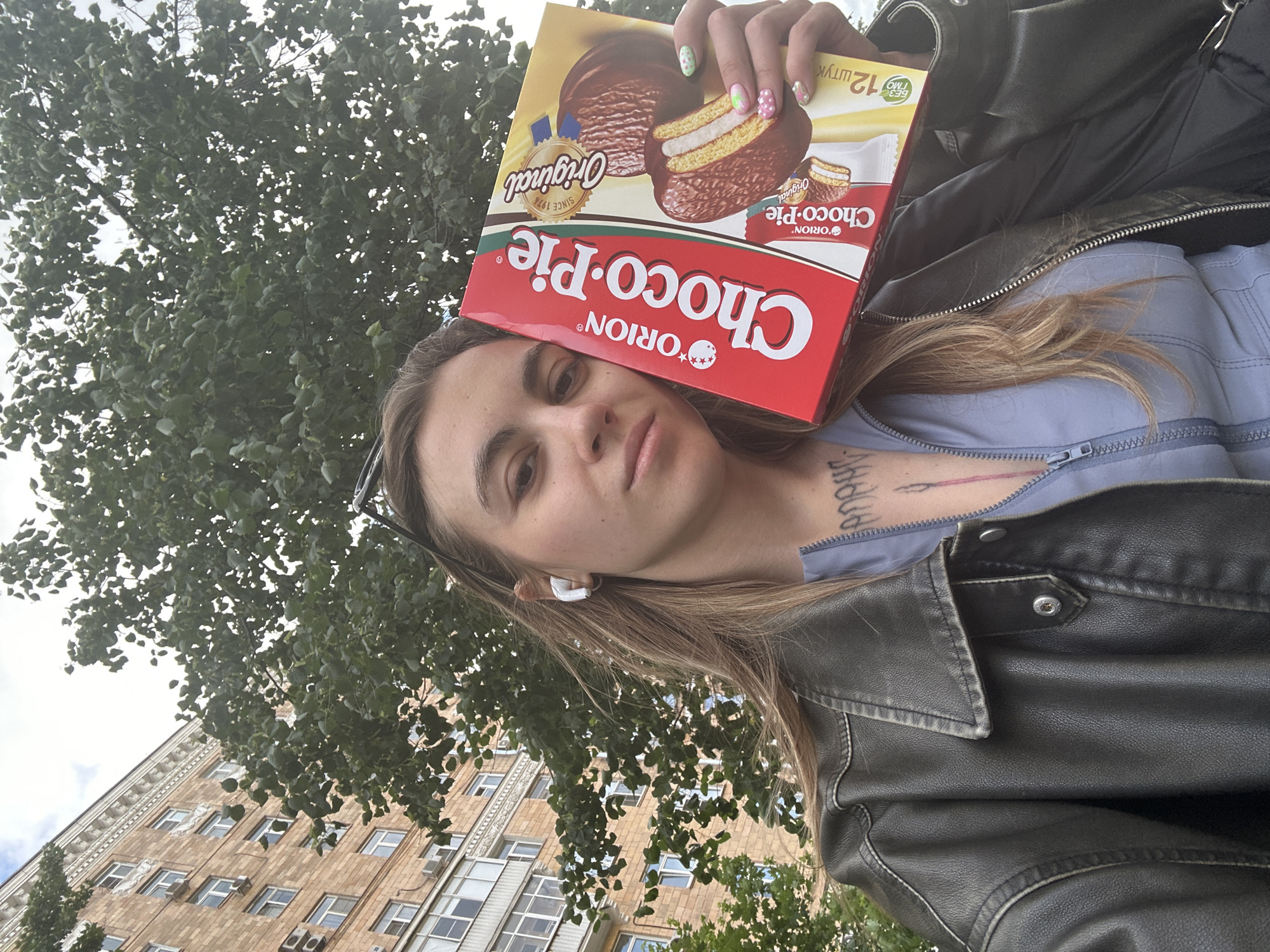 A young woman with long blonde hair, wearing a leather jacket and a blue zip-up top, poses for a selfie outdoors, holding a box of Orion Choco-Pie. She has earbuds in her ears and tattoos visible on her chest. Behind her, a multi-story brick building with visible air conditioning units and windows suggests an urban setting, likely in Moscow, Russia. The sky is overcast, and lush green trees are in the background.