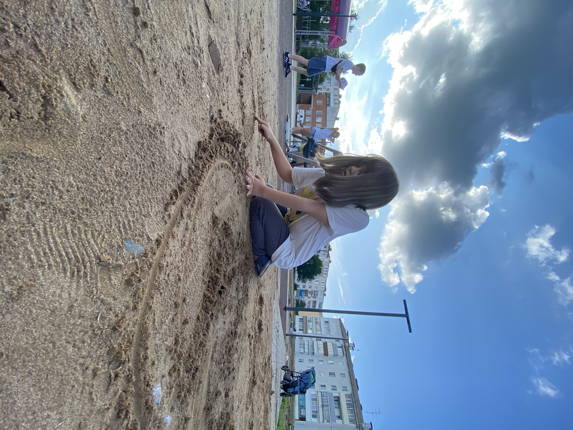 An outdoor, low-angle, full shot captures a young person in a white t-shirt and dark pants kneeling in a sandbox, drawing in the sand with their right index finger. The sandbox is textured with a pattern etched into the sand, possibly from a tire. In the background, on a playground, another young person in a white t-shirt and shorts is standing, holding a round object, and a young girl is standing with her hands behind her head. Further in the background, a blue baby stroller is parked next to a building. The sky is a bright blue with dramatic, dark clouds. The scene is set in a city in Noviy, Russia.