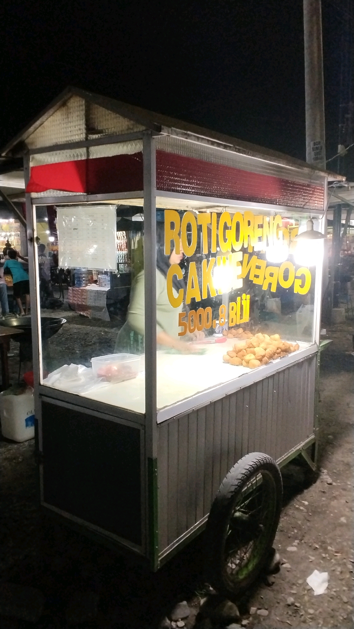 A street food cart at night in Canggu, Indonesia, displays a sign that reads "ROTIGORENG CAKE". The cart has a metal frame and is covered by a corrugated roof. A pile of fried food items sits on a counter inside the cart. A person wearing a headscarf and an olive-green shirt is visible behind the counter, preparing food. In the background, there are other stalls and people. The ground is dirt and rocks, and there is a wheel attached to the cart.