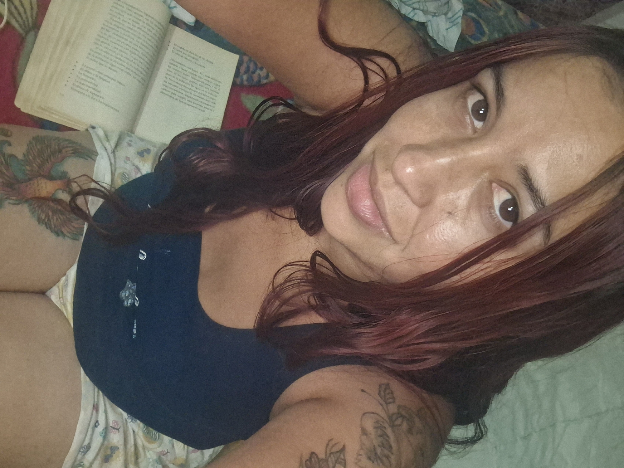 The image shows a woman with reddish-brown hair lying down. An open book is near her left arm. She has tattoos on her left thigh and right arm. The background includes a bed with patterned sheets. The location is not discernible from the image and cannot be determined to be Vallecito, Venezuela.