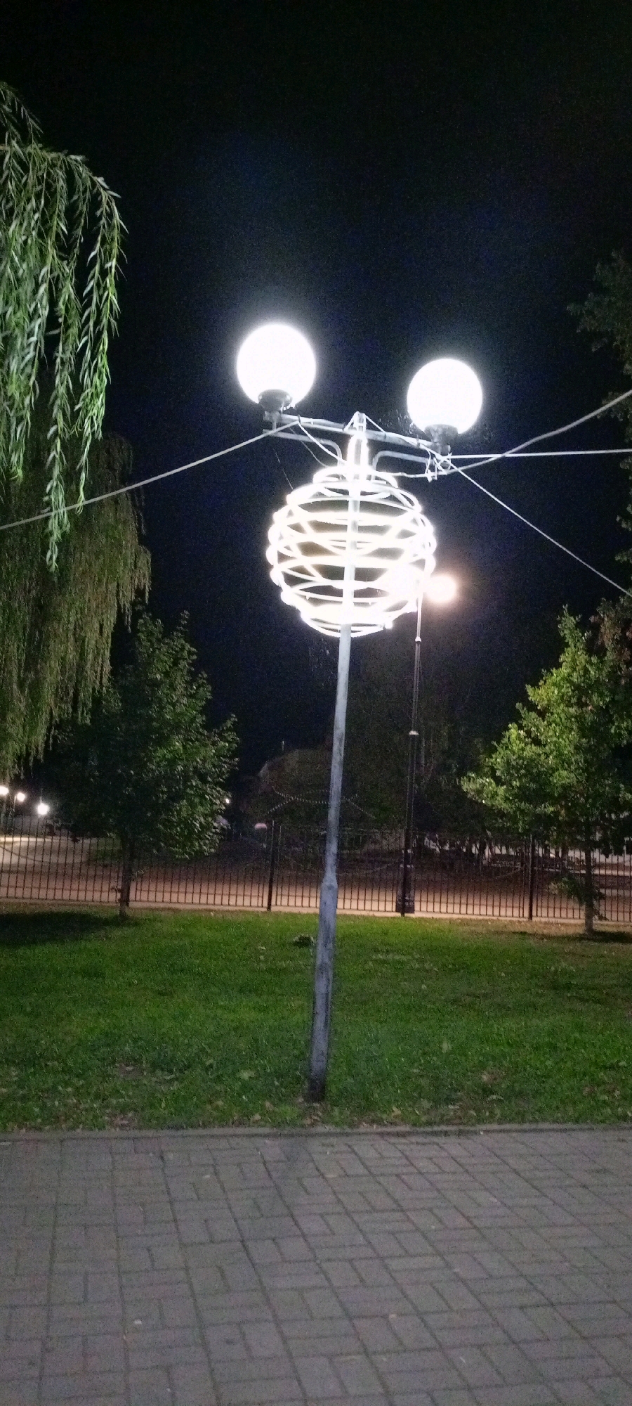The image shows a street lamp with a decorative globe in the city of Kirsanov, Russia, at night. The lamp post is made of metal and has two spherical light fixtures at the top. A large, spherical ornament made of white, illuminated wire is suspended from the lamp post. In the background, there is a grassy area with trees and a metal fence. The ground in the foreground is paved with grey interlocking bricks. The sky is dark and clear.