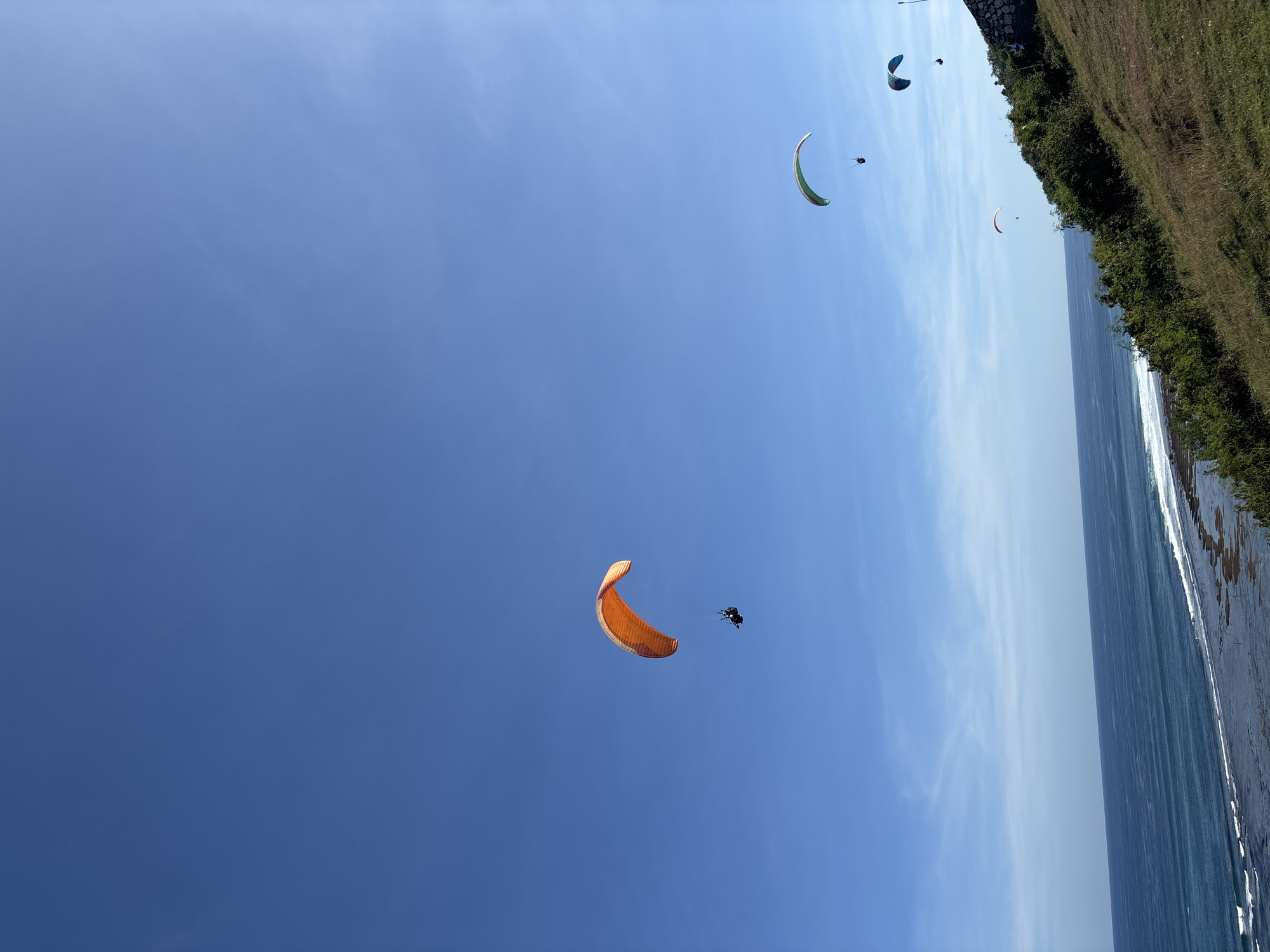 The image depicts several paragliders in the sky above the coastline of Kuta Selatan, Indonesia. The sky is predominantly blue with some wispy clouds. The coastline includes vegetation and a beach with waves. One paraglider in the foreground is orange.