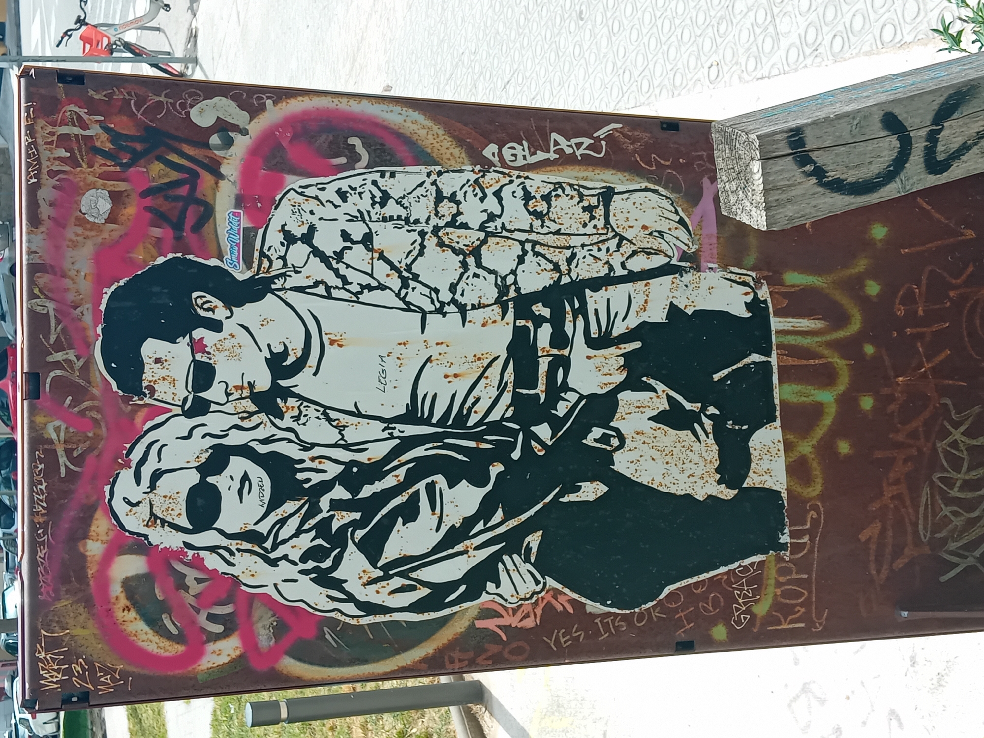 A graffiti-covered metal panel features a prominent black and white stencil-style artwork of two figures. The main figure is a man with dark hair and sunglasses, holding what appears to be a gun. He is embraced by, or embracing, a woman with long, wavy hair and sunglasses. Both figures are rendered in a stark, high-contrast style, with elements of rust showing through the white areas.
The metal surface itself is heavily rusted and covered in layers of additional graffiti tags in various colors, including bright pink, brown, green, and black. Some of these tags are partially obscured by the central artwork. In the top left corner, part of a bicycle is visible. A rough wooden beam extends from the top right edge of the panel. The background above the panel consists of a white, textured wall and some green foliage. The bright, even lighting suggests it is daytime.
Visible text includes: "Snuts Widdet" on a sticker near the top figure; "AMER. F.!" on the left edge; "LEGIA" on the man's arm; "MDZEN" on the woman's hair; "YES. ITS OK", "GREAC", and "KOPOS" below the figures; and "QLARÍ" (possibly "POLARÍ") above the figures on the right. The scene captures a vibrant piece of street art against an urban backdrop in Barcelona, Spain.