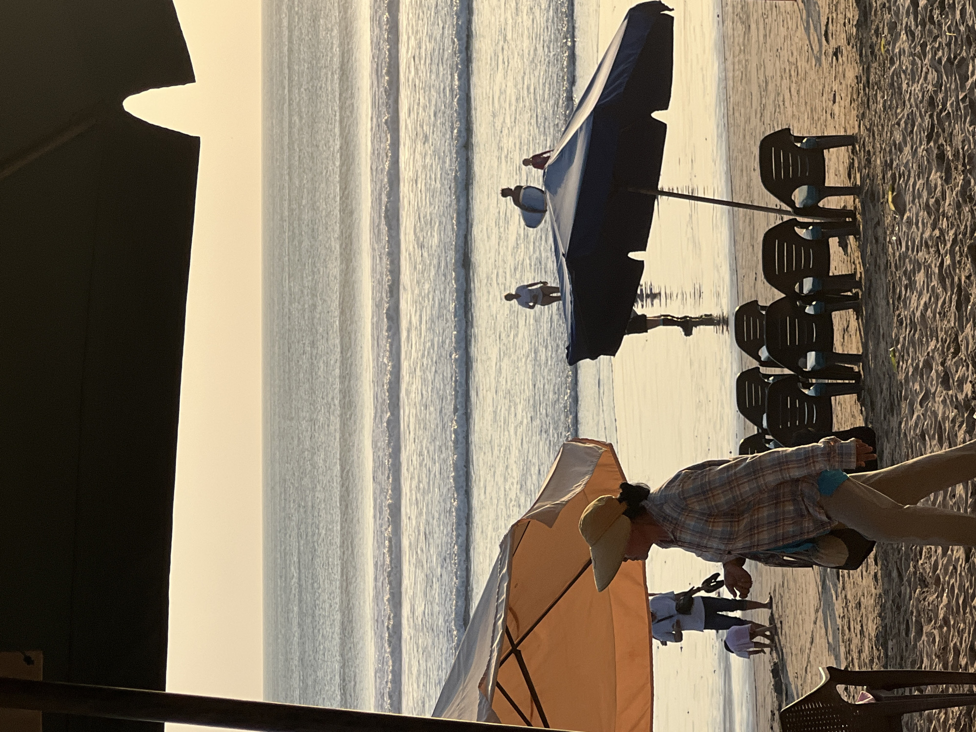 This image depicts a sunset scene on Kuta Beach, Indonesia. In the foreground, several dark plastic beach chairs are arranged under a large, dark blue umbrella. To the left, a woman wearing a wide-brimmed hat and a plaid shirt walks across the sand, carrying a bag over her shoulder. Behind her, an orange umbrella shades a person standing. In the middle ground, several people are visible in the shallow ocean water, with one person carrying a surfboard. The ocean exhibits gentle waves that are catching the warm light of the setting sun. The sky is a soft, hazy orange and yellow, typical of a tropical sunset. The overall atmosphere is relaxed and tranquil.