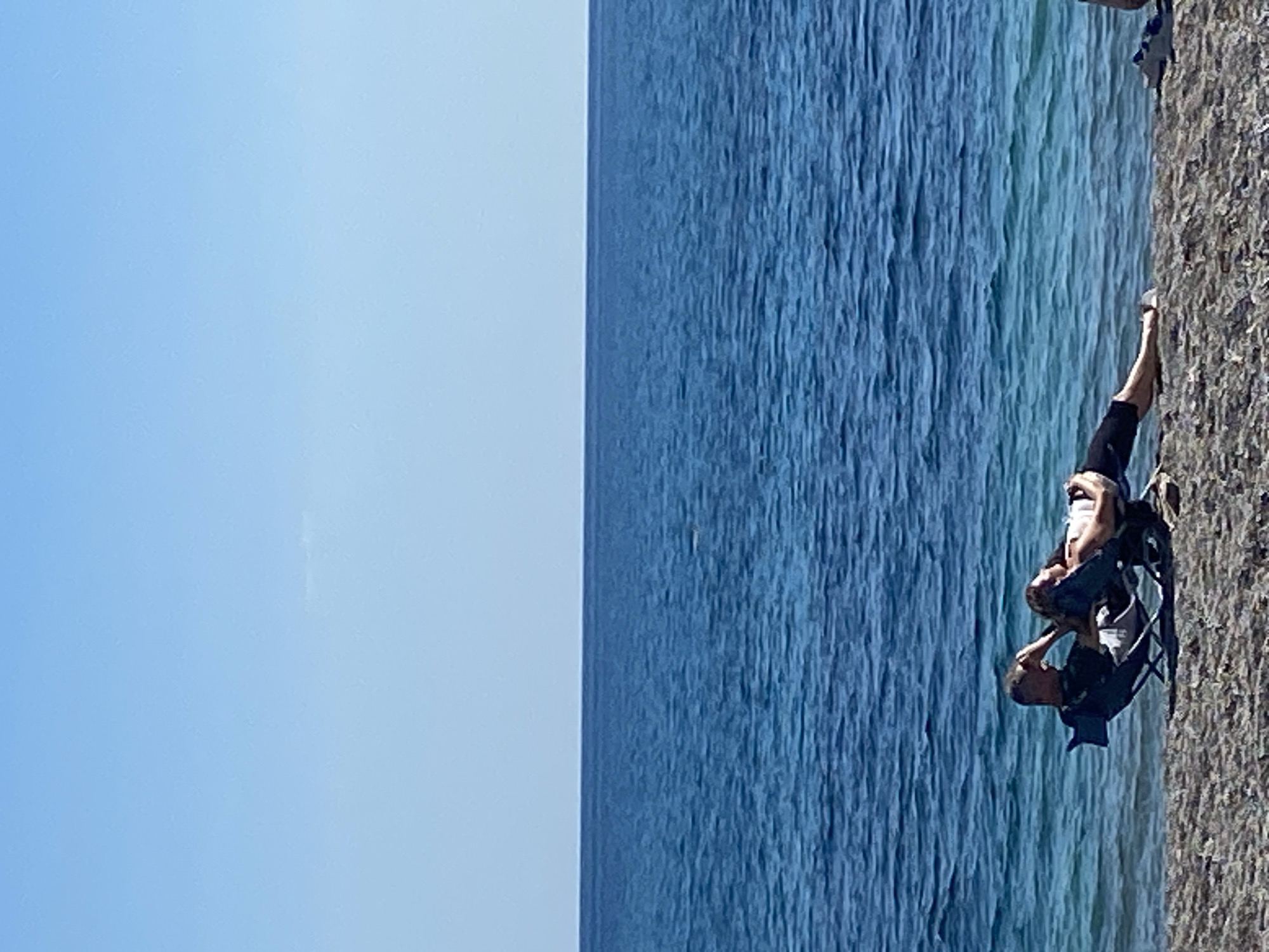 The image depicts a landscape with a body of water and a person sitting in a chair on the adjacent shore. A clear blue sky is visible on the left. The water is dark blue with visible texture indicating waves. On the right, the shore appears to be rocky or sandy. A person is seated in a foldable chair on the shore, facing the water. The location is likely in Prijevor, Montenegro, based on the prompt.