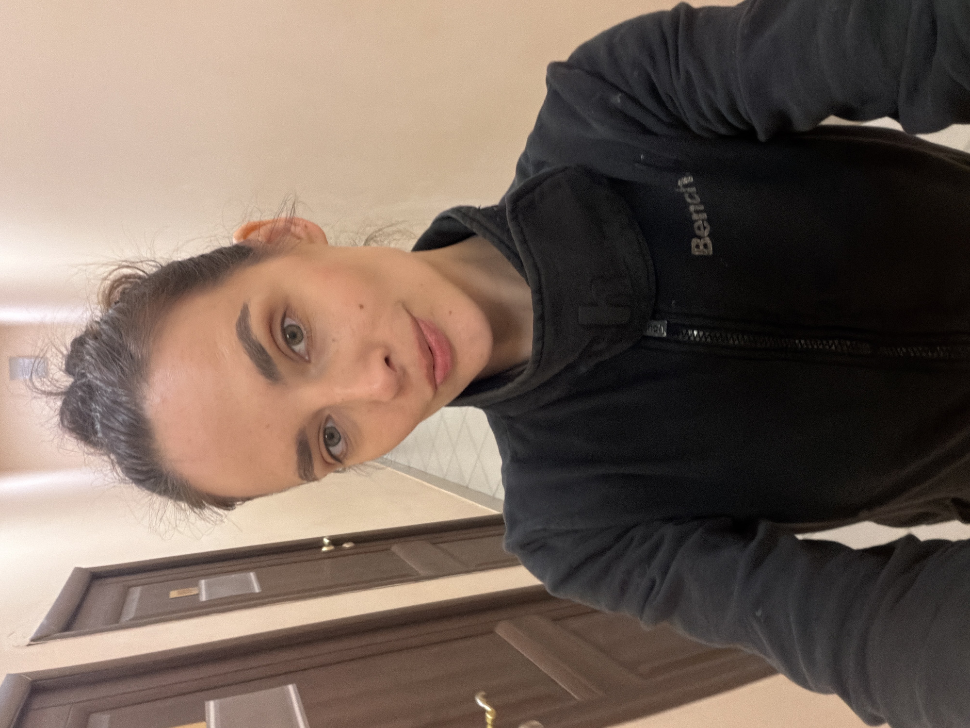 A woman with dark hair pulled up in a bun takes a selfie in a narrow hallway. She is wearing a black zip-up jacket with the word "Bench" visible on the chest. The hallway has beige walls and two closed wooden doors on the left side. The floor is tiled. The image does not provide any specific location markers that would identify it as being in Ufa, Russia.