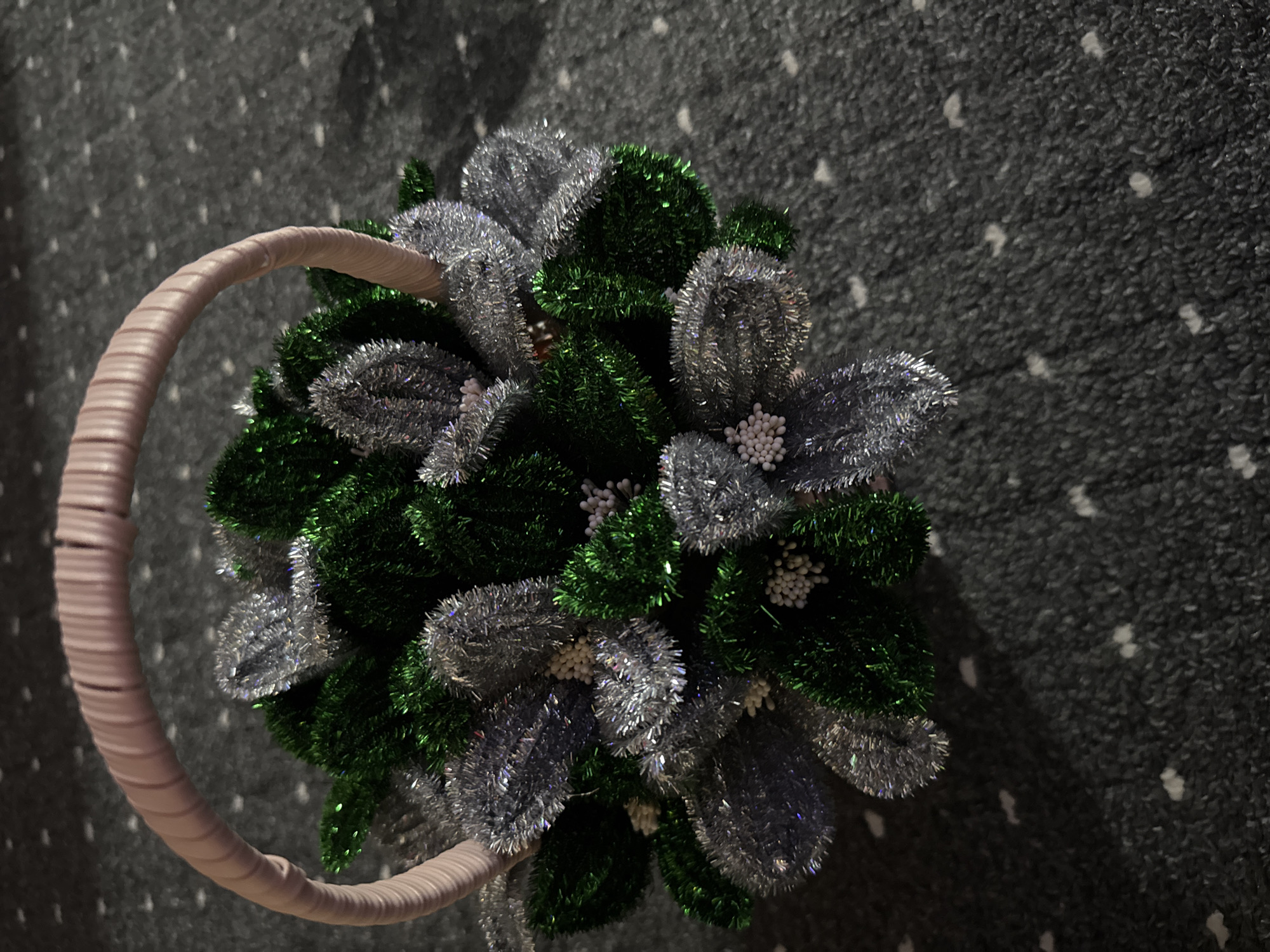 A close-up, high-angle shot shows a small, decorative basket filled with artificial greenery and silver tinsel foliage, set against a dark, textured carpet. The basket has a light pink, ribbed handle arching over the top. The foliage is arranged in a dense, circular pattern, with some small white bead-like embellishments scattered among the leaves. The carpet has a subtle pattern of lighter specks. The image is well-lit, highlighting the shimmering textures of the tinsel. The specific location of Chardara, Uzbekistan, cannot be determined from the visual information provided in the image.