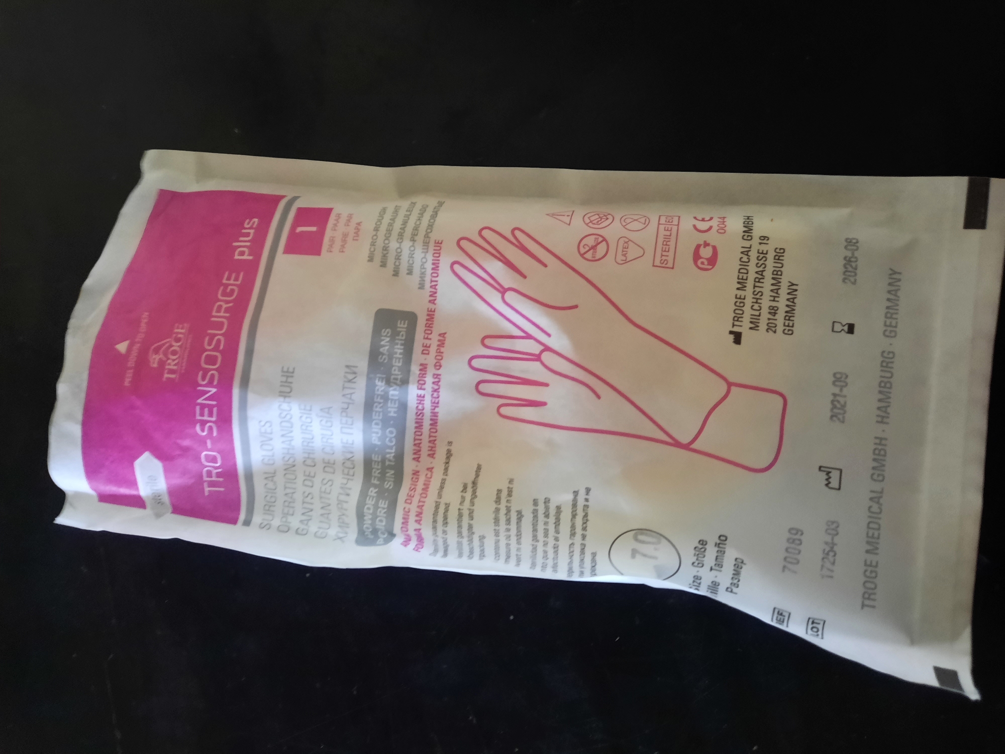 A package of surgical gloves, labeled "TRO-SENSOSURGE plus," is displayed against a dark, flat surface, likely a table. The package is predominantly white with pink accents, featuring text in multiple languages, including English, German, French, Spanish, and Russian. The gloves inside are depicted in outline, a stylized pink image of a pair of hands with fingers extended. Several certifications and information labels are visible on the package, including "STERILE," "LATEX," "CE 0044," and a barcode-like symbol. The size of the gloves is indicated as "7.0." Manufacturing details such as "TROGE MEDICAL GMBH," "MILCHSTRASSE 19," "20148 HAMBURG," and "GERMANY" are present, along with a lot number "70089," a manufacturing date of "2021-09," and an expiration date of "2026-08." The image is a close-up, focusing on the product packaging. There is no discernible information in the image that would place this scene in Lhokseumawe, Indonesia.