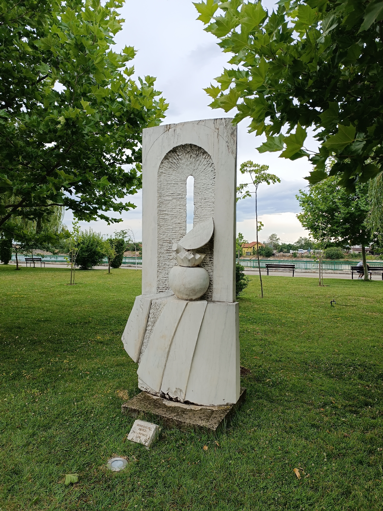 The image depicts a large, abstract white stone sculpture situated in a grassy park. The sculpture has a rectangular base and features a curved, arched opening at the top. Below the opening, there is a spherical element with a flat, disc-like shape resting on top of it, all rendered in stone. The sculpture is positioned on a concrete base, with a small stone plaque at its foot inscribed with text, possibly the artist's name and year.
The park setting includes well-maintained green grass, scattered trees, and a body of water in the background, likely the Jiu River, with a paved walkway and benches along its bank. In the distance, buildings and more trees are visible. The sky is overcast. This sculpture is part of Târgu Jiu's public art ensemble, created by Constantin Brâncuși, and is known as "The Table of Silence" or "Masa Tăcerii" in Romanian. The sculpture itself is a representation of a circular table with accompanying stone stools, though this particular sculpture shown is not the classic "Table of Silence" ensemble. It is located near the Jiu River in Târgu Jiu, Romania.