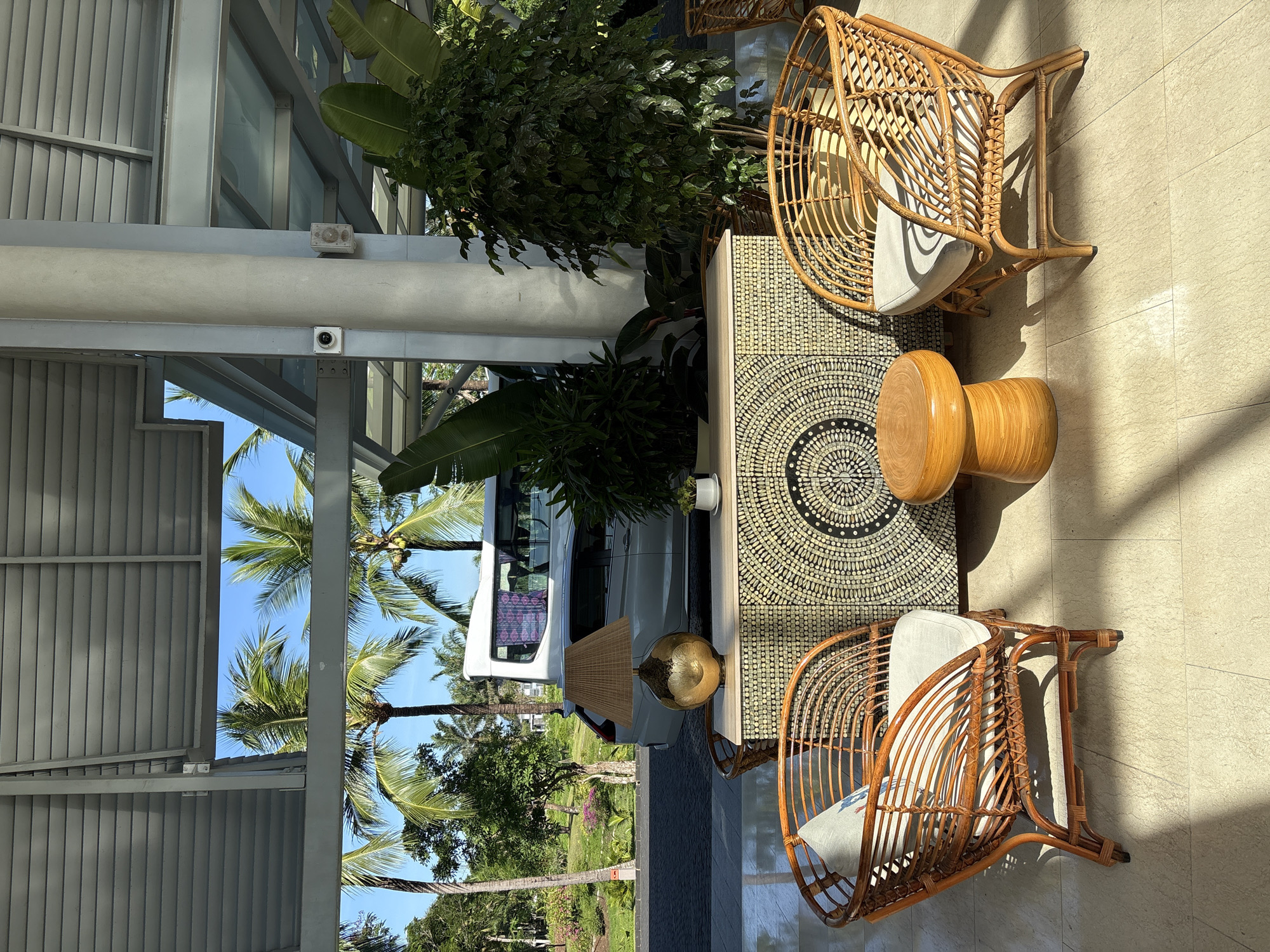 The image shows an outdoor seating area, likely a hotel lobby or restaurant, in Đà Nẵng, Vietnam. Two rattan armchairs with light-colored cushions are positioned on either side of a decorative cabinet. A small, round wooden stool sits in front of the cabinet. The cabinet itself is adorned with a circular, mosaic-like pattern of light-colored circular objects on a dark background. A lamp with a woven shade and a golden, bulbous base sits on the cabinet. Behind the cabinet, a large green plant is visible. In the background, palm trees and other lush vegetation are evident, under a bright blue sky. A white van is parked on a paved area. The area appears to be covered by a roof structure supported by white columns. Sunlight casts dappled shadows across the floor.
