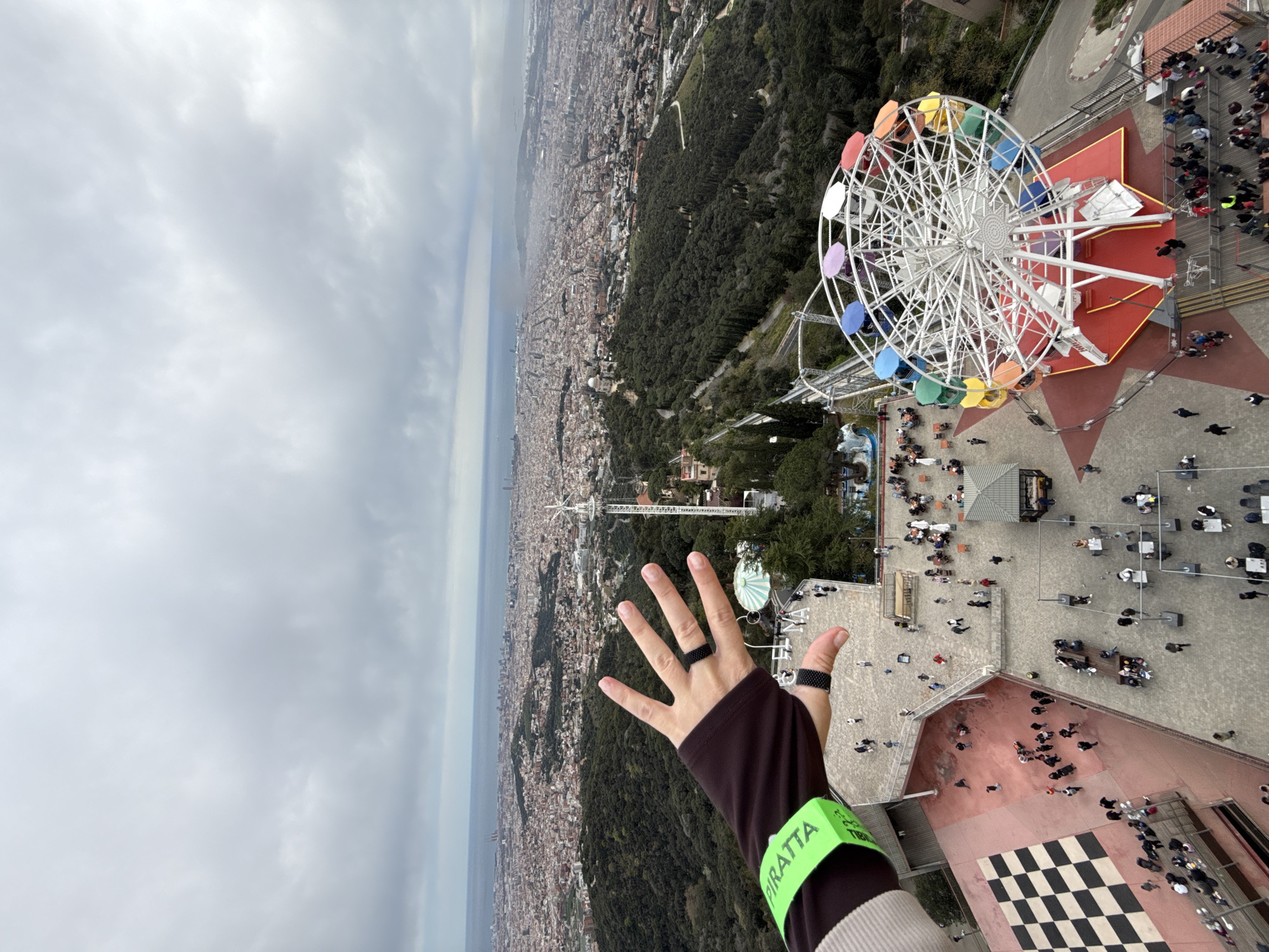 A vertically-oriented, high-angle photograph reveals a scenic overlook in Barcelona, Spain, likely taken from Tibidabo Mountain, offering panoramic views. The skyline of Barcelona stretches to the horizon where the sea meets a cloudy sky. A dense urban landscape transitions into a forested area leading up to the mountain.
In the foreground on the right, a Ferris wheel with rainbow-colored cabins stands prominently. Nearby, groups of people are clustered, suggesting an amusement park or observation point. A hand with rings on the fingers is held up in the lower center of the frame, wearing a wristband labeled "TIBIDABO PIRATTA" in bold, green lettering. The wristband is part of a brown and cream colored long sleeve shirt.
The composition captures a sense of altitude and the scale of the city below, along with the recreational atmosphere of the mountain top.