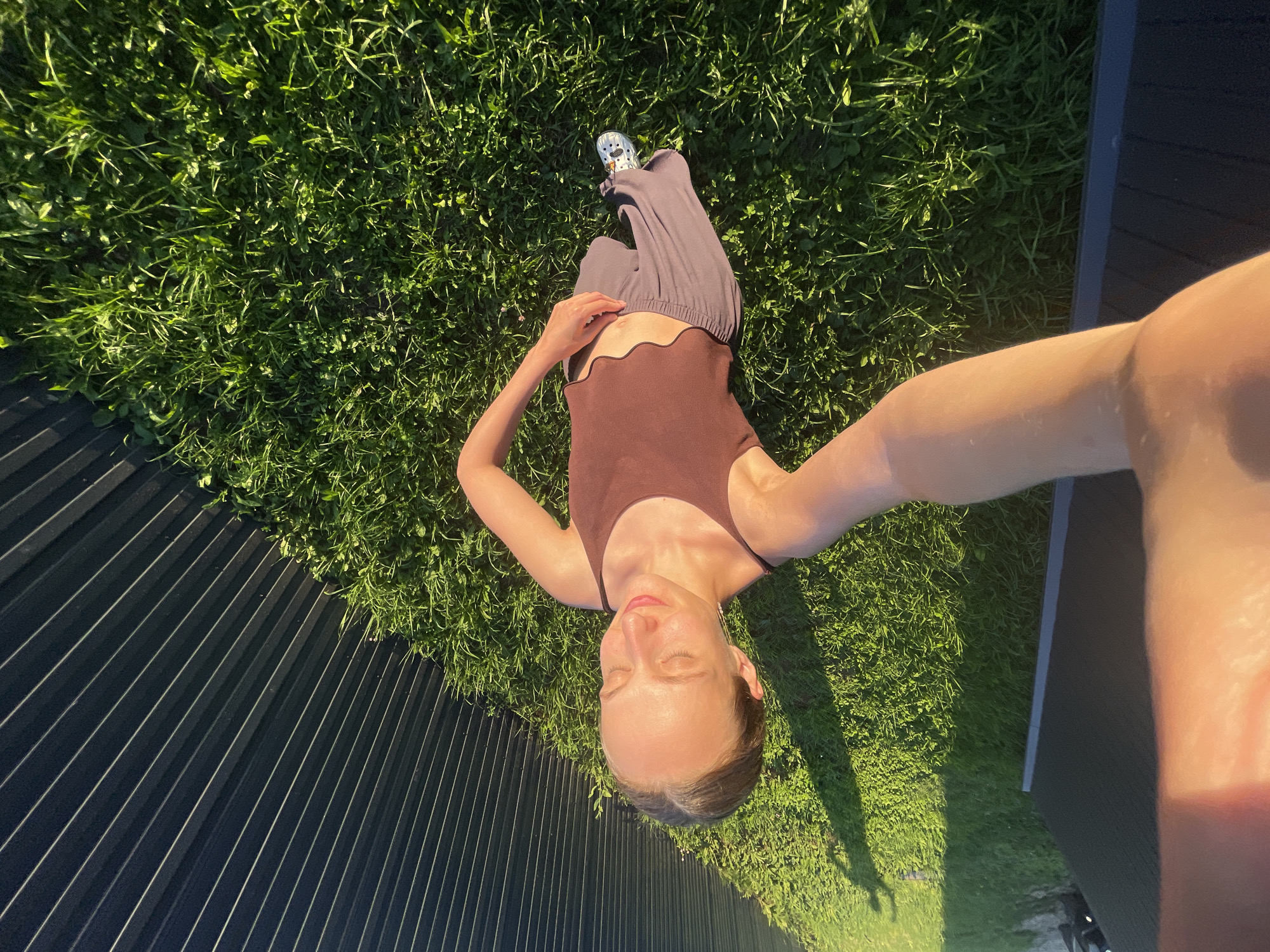 A young woman is standing in a grassy area next to a dark grey corrugated metal fence. She is taking a selfie with her left arm extended, holding the camera. She is wearing a brown tank top and grey sweatpants, with white and silver sneakers on her feet. Her eyes are closed, and she is smiling slightly. The sun is casting a long shadow on the grass from the fence and from an unseen object to the left of the frame. The location is outdoors in a residential area, possibly a backyard, in the city of Podshivalovo, Russia.