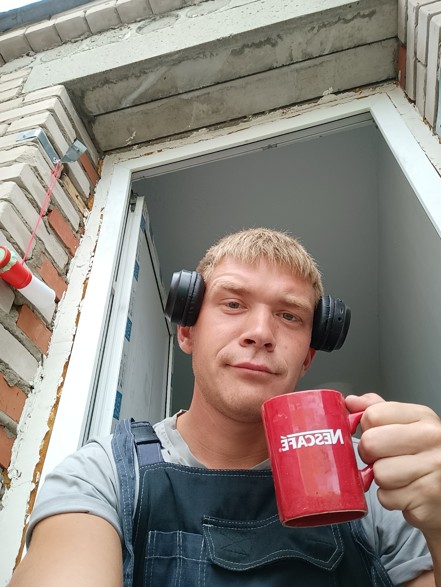 In a close-up, low-angle selfie, a light-skinned male with short blonde hair and light eyes is pictured outdoors, holding a red Nescafe mug in his right hand and wearing black over-ear headphones. He is dressed in a grey t-shirt and blue overalls. The background shows a brick wall and the frame of an open window or doorway. A red and white plastic object with a green cap is attached to the brick wall by a metal bracket and a red string. The man appears to be looking directly at the camera.