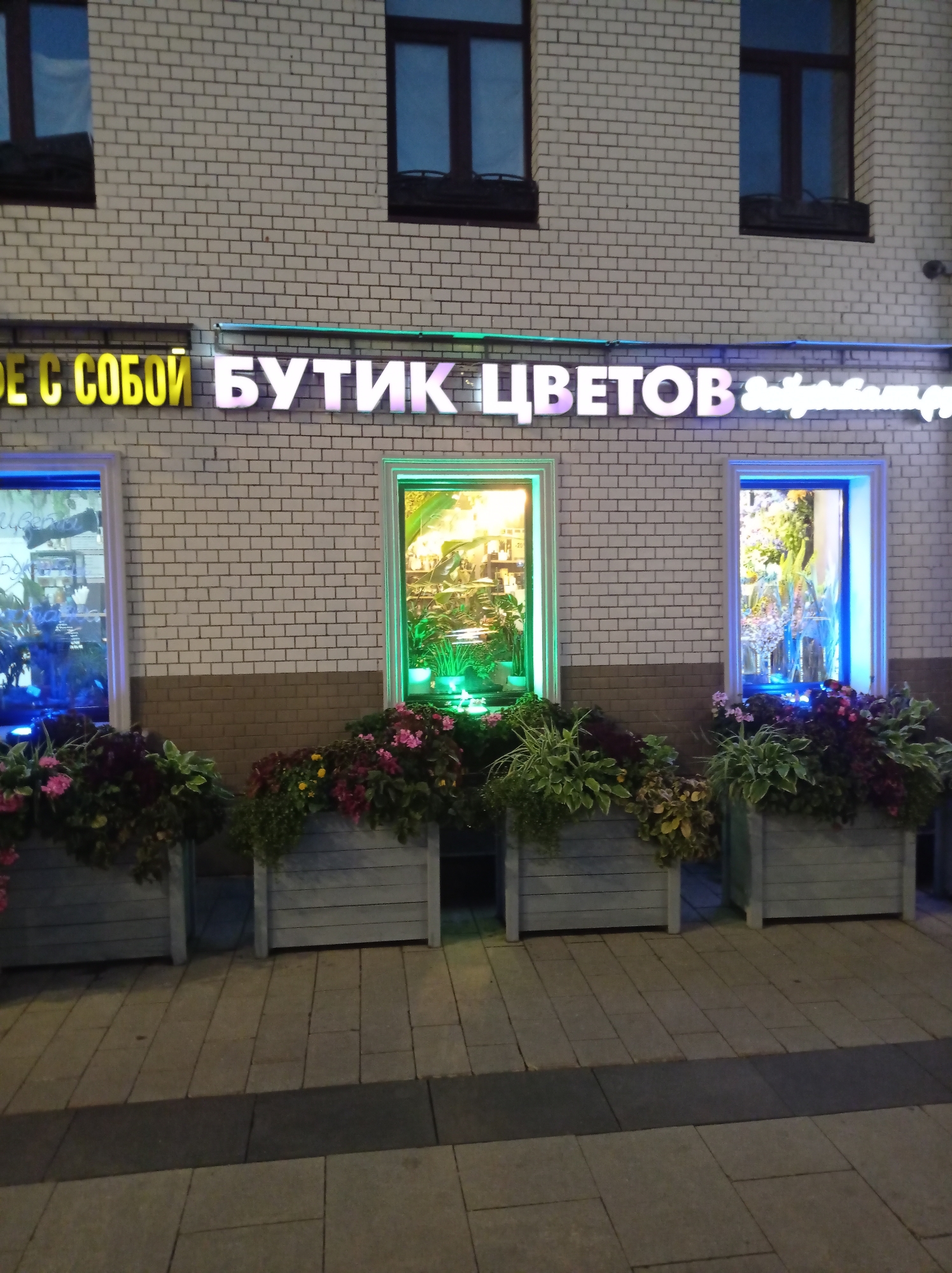 This is a nighttime outdoor photograph of a flower shop located in Moscow, Russia. The shop is situated in a building with a white brick facade and features three illuminated windows and a prominent illuminated sign. The sign reads "БУТИК ЦВЕТОВ" which translates to "Flower Boutique" in English, with additional Cyrillic text to the right. The windows display arrangements of plants and flowers, with colored lights illuminating the left and right windows in blue and the central window in green. In front of the building, there are several large rectangular planters containing a variety of flowering plants and foliage. The ground in front of the planters is paved with light-colored square and rectangular stones. The overall scene suggests a well-maintained and inviting retail establishment.