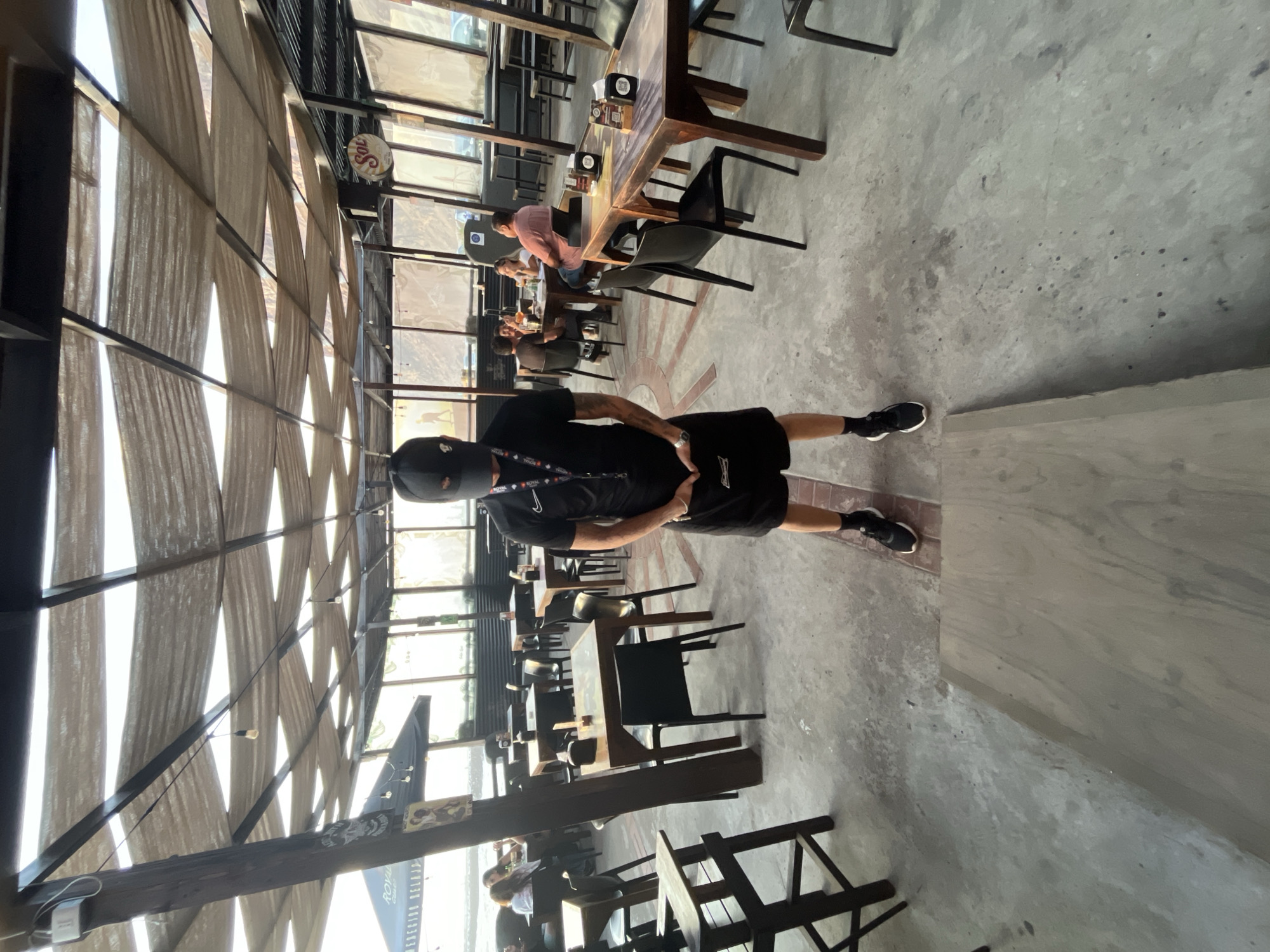 A man wearing a black ski mask, black shirt, black shorts, and black shoes stands in a restaurant or bar in Arica, Chile. The building is open-air with a high ceiling supported by metal beams and covered with white fabric. There are multiple tables and chairs, some of which are occupied by other patrons. The floor is concrete. A sign with "Sol" is visible.