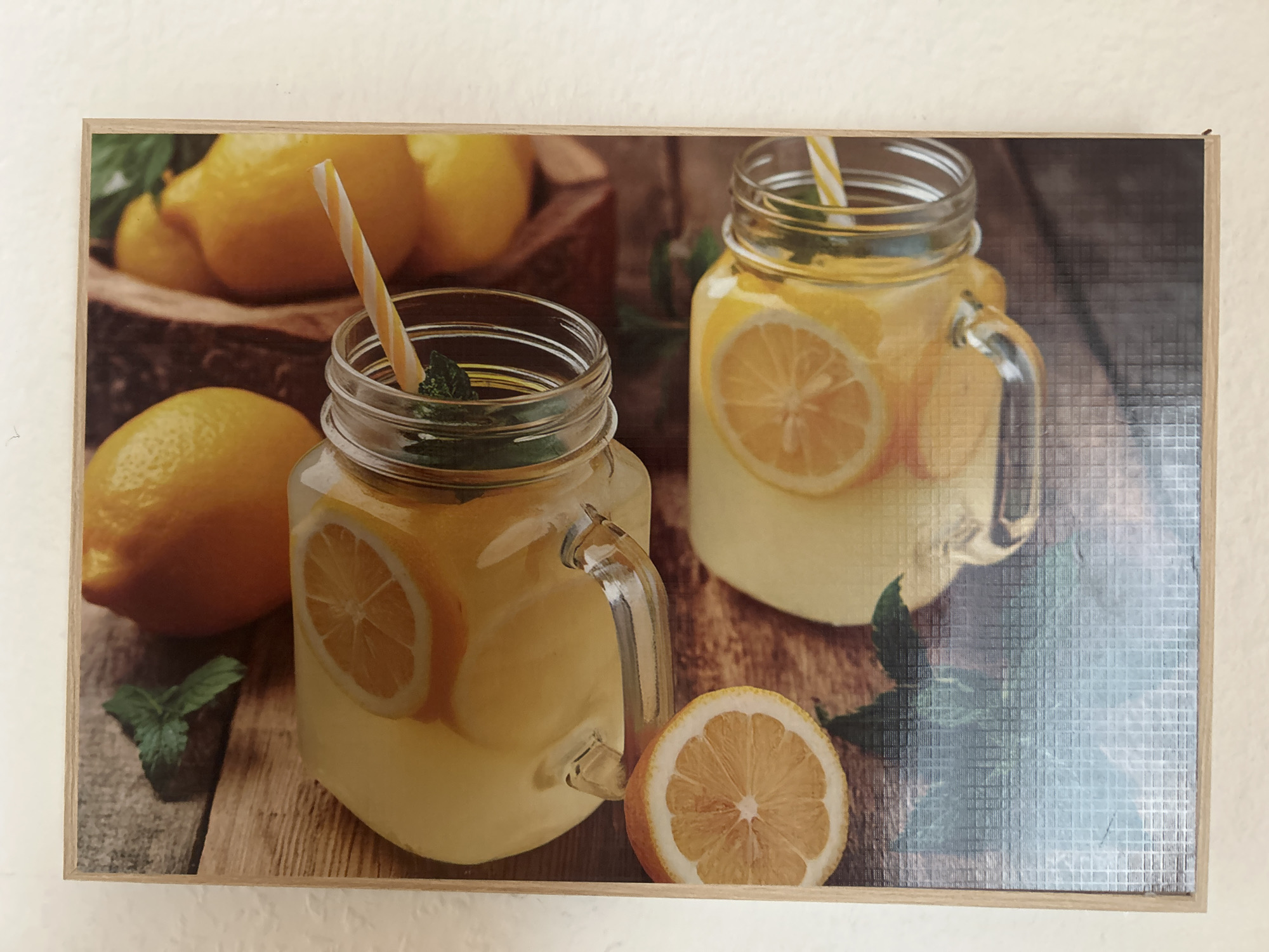 This is a photograph of a framed artwork depicting two glasses of lemonade with lemons and mint. The artwork is mounted on a wall. The lemonade is in mason jar glasses and appears to be refreshing, with slices of lemon and mint leaves visible. The background consists of lemons and a wooden surface. The overall scene is bright and inviting. There is no information about the location in Detmold, Germany.