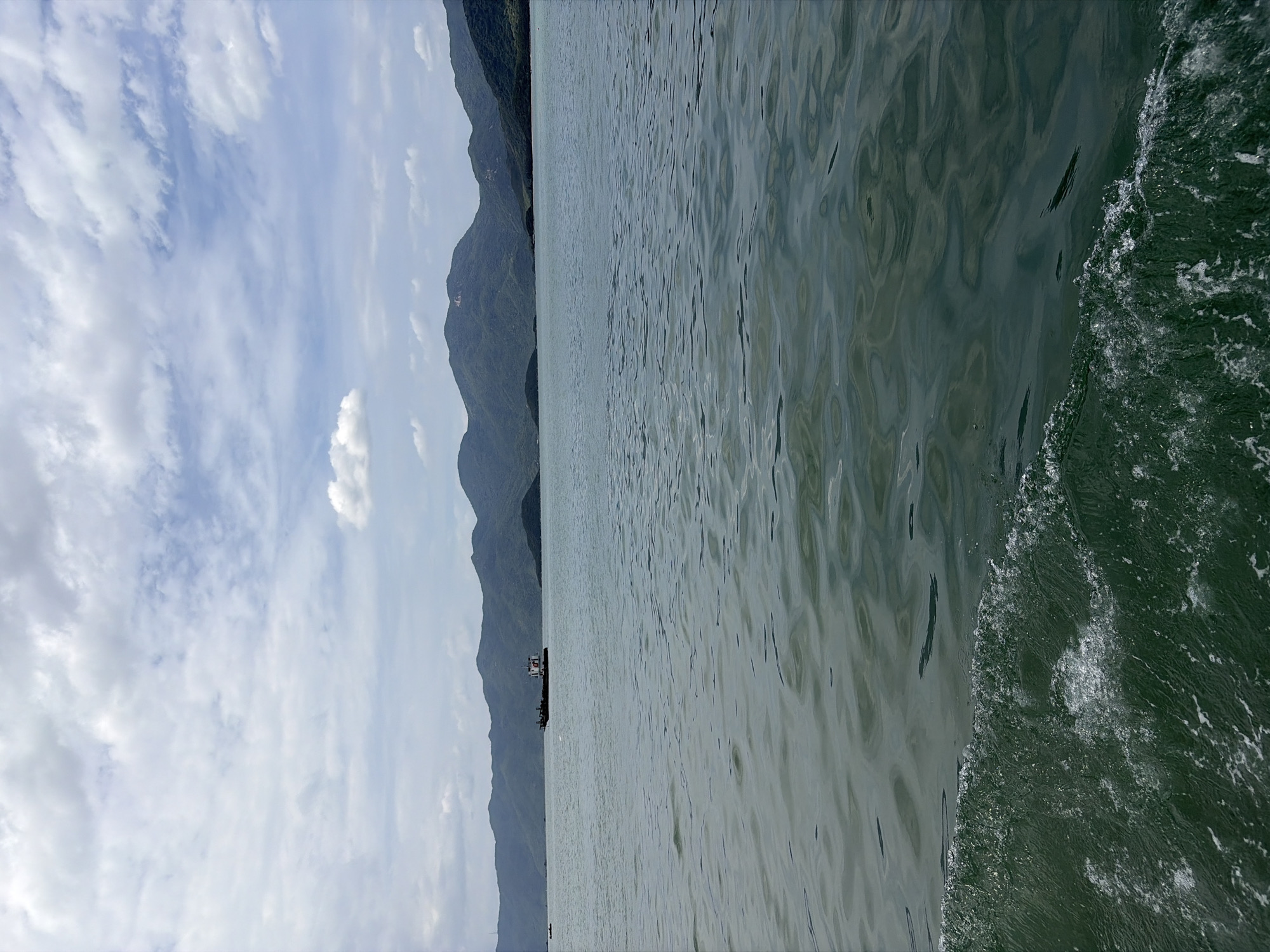 The image depicts a view from a body of water, likely a sea or large lake, with a mountainous coastline in the background. In the middle ground, a dark-colored barge with a white and red structure on its deck is visible. The water in the foreground shows a white foamy wake, suggesting movement. The sky is partly cloudy with patches of blue visible. The mountainous terrain is covered in green vegetation. The scene is likely located in the coastal region of Ninh Ích, Vietnam, given the landscape and prevalent nautical activity in such areas.