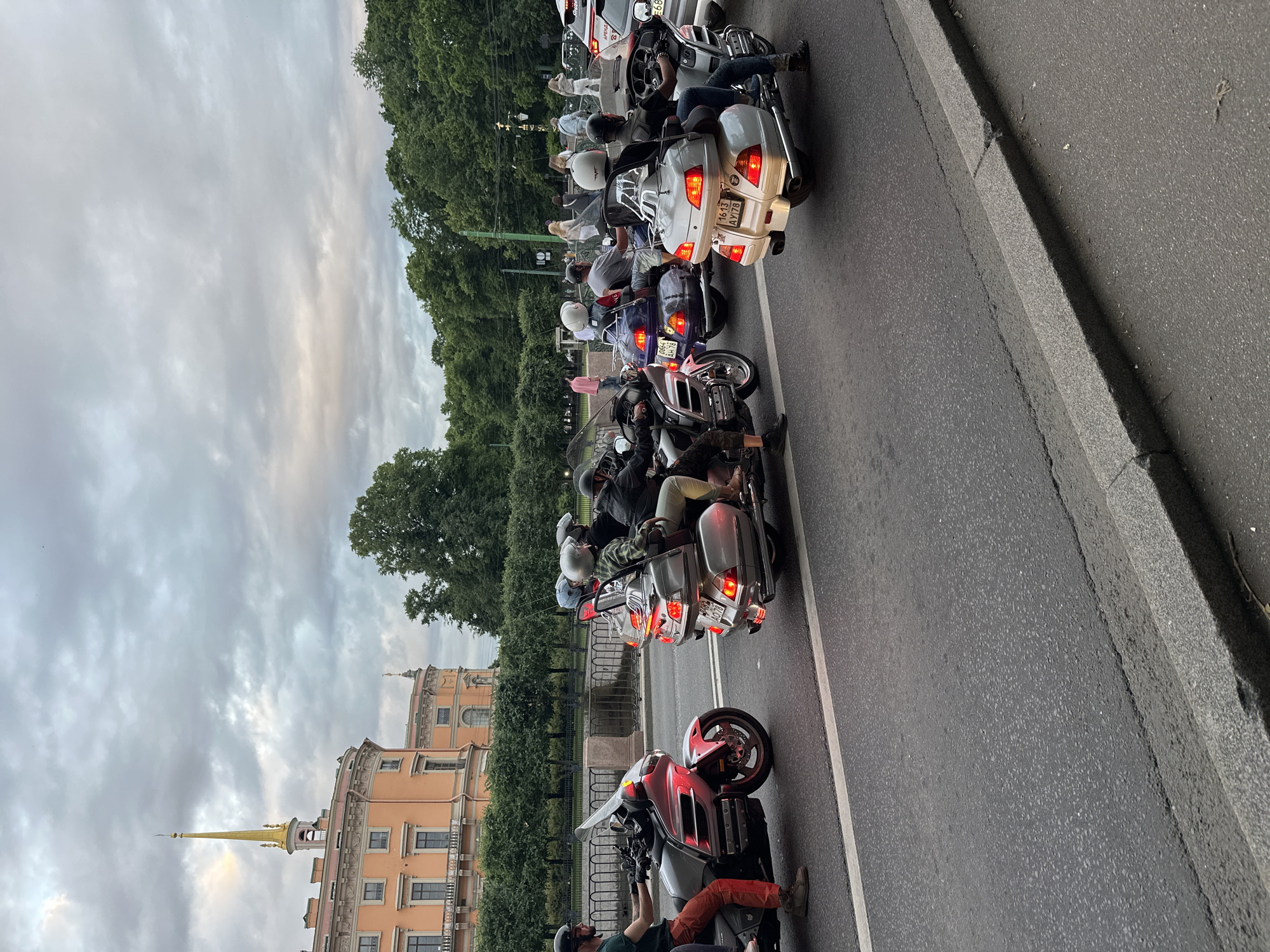 A group of motorcyclists are riding on a road in front of a large, pastel-colored building with a golden spire. The building appears to be a historical landmark, likely in Saint Petersburg, Russia, given the architectural style and the presence of trees lining a canal or river behind a fence. The sky is overcast with dramatic clouds. The motorcyclists are all wearing helmets and riding large touring motorcycles. Some are in the foreground, while others are further back, creating a sense of depth. The road is asphalt, with a white lane marker visible. In the background, there are more trees and a few pedestrians walking. The overall scene depicts a group ride through a scenic, urban environment.