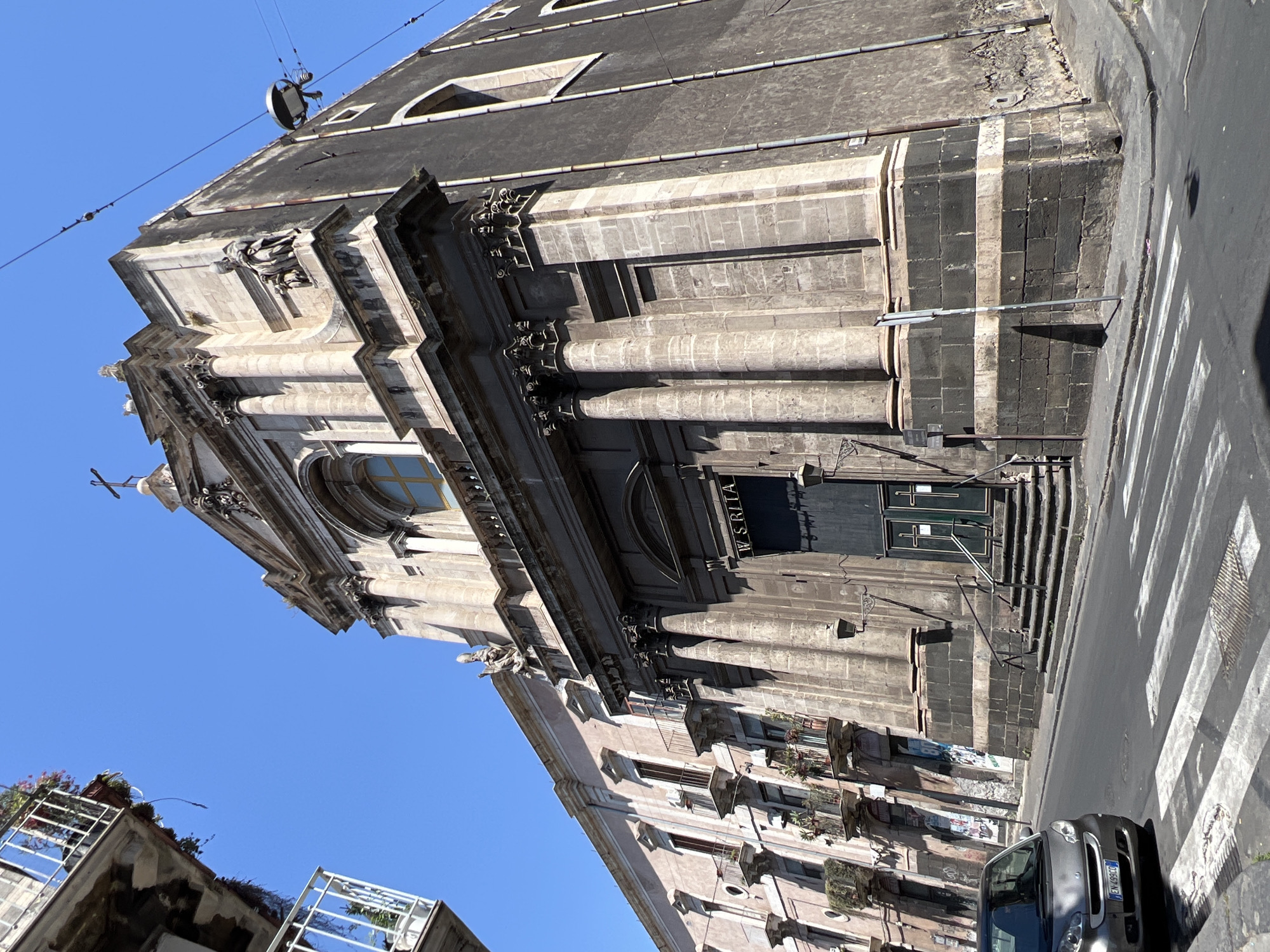 The image depicts a street view of a church in Catania, Italy, on a clear, sunny day. The church, with its imposing stone facade featuring classical columns and architectural details, dominates the right side of the frame. Above the entrance, the words "W. SRITA" are visible. Adjacent to the church, to the left, is a building with multiple balconies adorned with potted plants. A small, gray car is parked on the street in the foreground, with a pedestrian crossing marked by white stripes. Electrical wires are strung across the sky, and a street lamp is visible attached to the church's exterior. The overall impression is of an urban setting, typical of historical Italian cities.