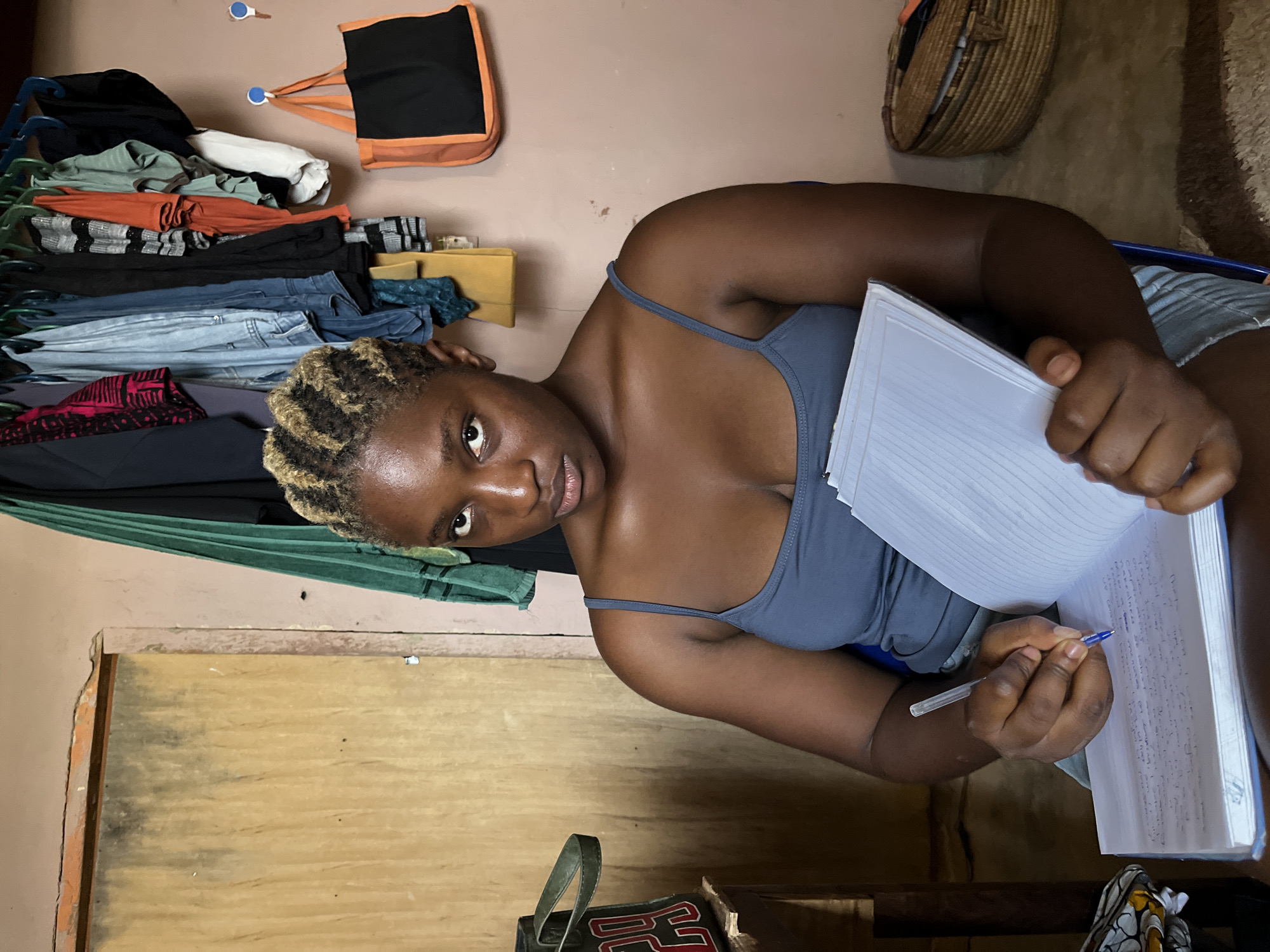 The image shows a young Black woman with blonde-streaked hair sitting and writing in a notebook. She is wearing a grey tank top. The setting appears to be an indoor room with a pinkish-brown wall and wooden furniture. To the left of the woman, there is a closet or shelf filled with folded clothes, including jeans and patterned fabrics. An orange and black bag hangs on the wall above the clothes. To the right of the woman, a woven basket is visible on a shelf. The lighting suggests it could be daytime. The woman's expression is neutral as she focuses on her writing. There is no visible text in the image. The overall impression is of a casual, domestic scene.