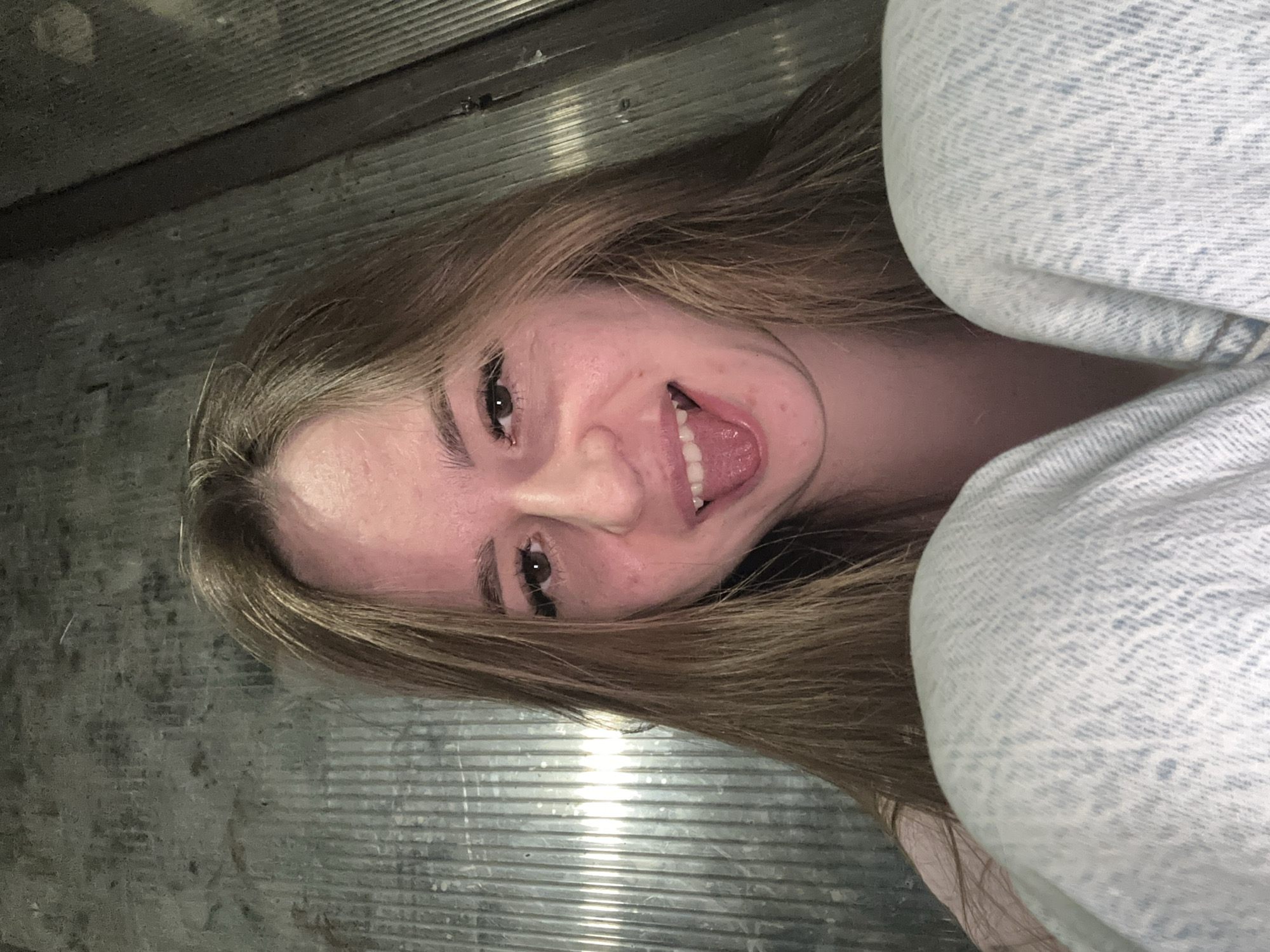 The image features a young woman posing for a selfie, her head tilted, suggesting she is lying down or looking up from a low angle. She has long, light brown hair, fair skin, and dark eyes. Her expression is playful, with a wide smile and her tongue slightly extended.
She is wearing light-colored, possibly acid-wash or faded, denim clothing, which is visible on the right side of the frame, covering part of her body or leg.
The background consists of a dark, textured surface with prominent horizontal ridges, resembling corrugated metal or a similar industrial material. There are reflections of light on this surface, indicating a light source, though the overall lighting appears somewhat dim. The image is oriented sideways, further suggesting the angle at which it was taken. There is no visible text in the image.