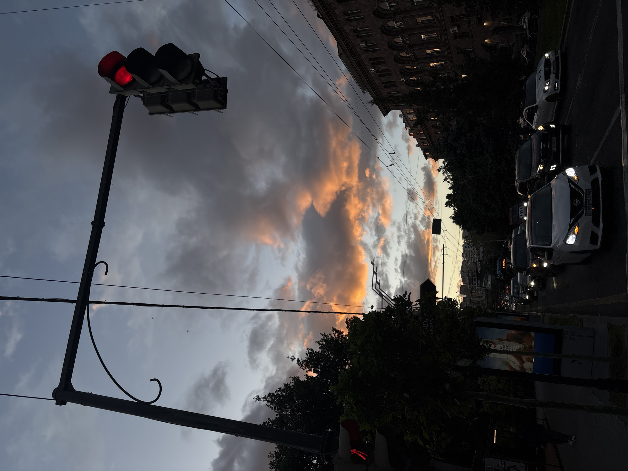 The image shows a street scene in Yerevan, Armenia at dusk. The camera is tilted, placing a red traffic light in the upper left corner, and a line of cars on the right side. A building with multiple stories and arched windows is visible above the cars. Power lines stretch across the sky. The sky is filled with dark clouds illuminated by orange light. Trees frame the bottom half of the view. Street-level advertisements are partially visible at the bottom.
