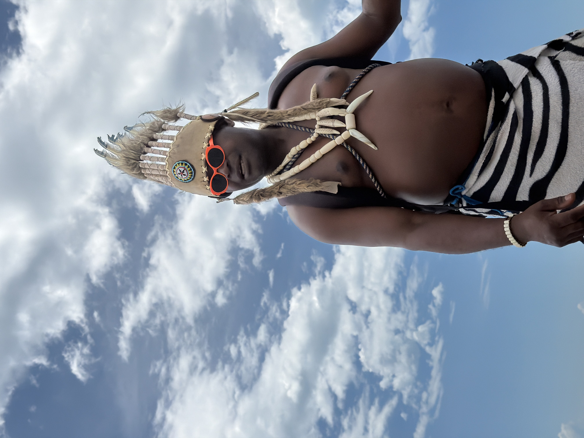 An adult male is photographed outdoors against a sky with scattered clouds. The man is wearing an elaborate headdress adorned with feathers and a beaded circular emblem. He has bright orange sunglasses on. Around his neck, he wears a necklace made of bone or shell pieces. He has a black and white striped garment wrapped around his waist and a bracelet of similar beads on his left wrist. The man is looking to his left with his mouth slightly open. The image does not provide any information about the location being Sochi, Russia.